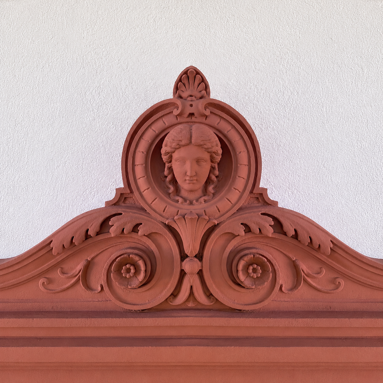 Eine kunstvolle Holzdekoration mit einem weiblichen Portrait in der Mitte, umgeben von filigranen ornamentalen Mustern auf einer weißen Wand.