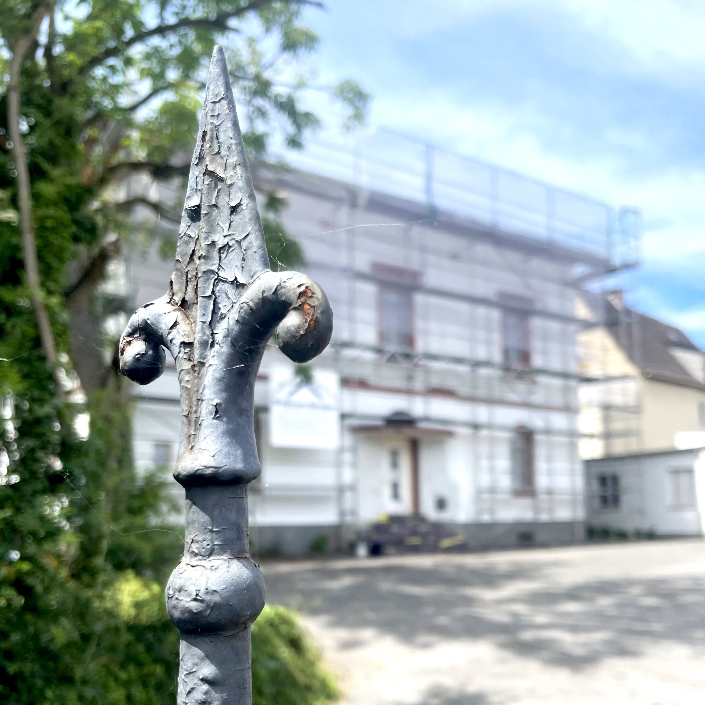 Ein metallener Zaun mit wetterfester, silbergrauer Lackierung, der an einer Seite einer Straße steht. Im Hintergrund sind grüne Bäume, ein blauer Himmel mit wenigen Wolken und eine Straße mit einer Brücke oder einem Überweg sichtbar.