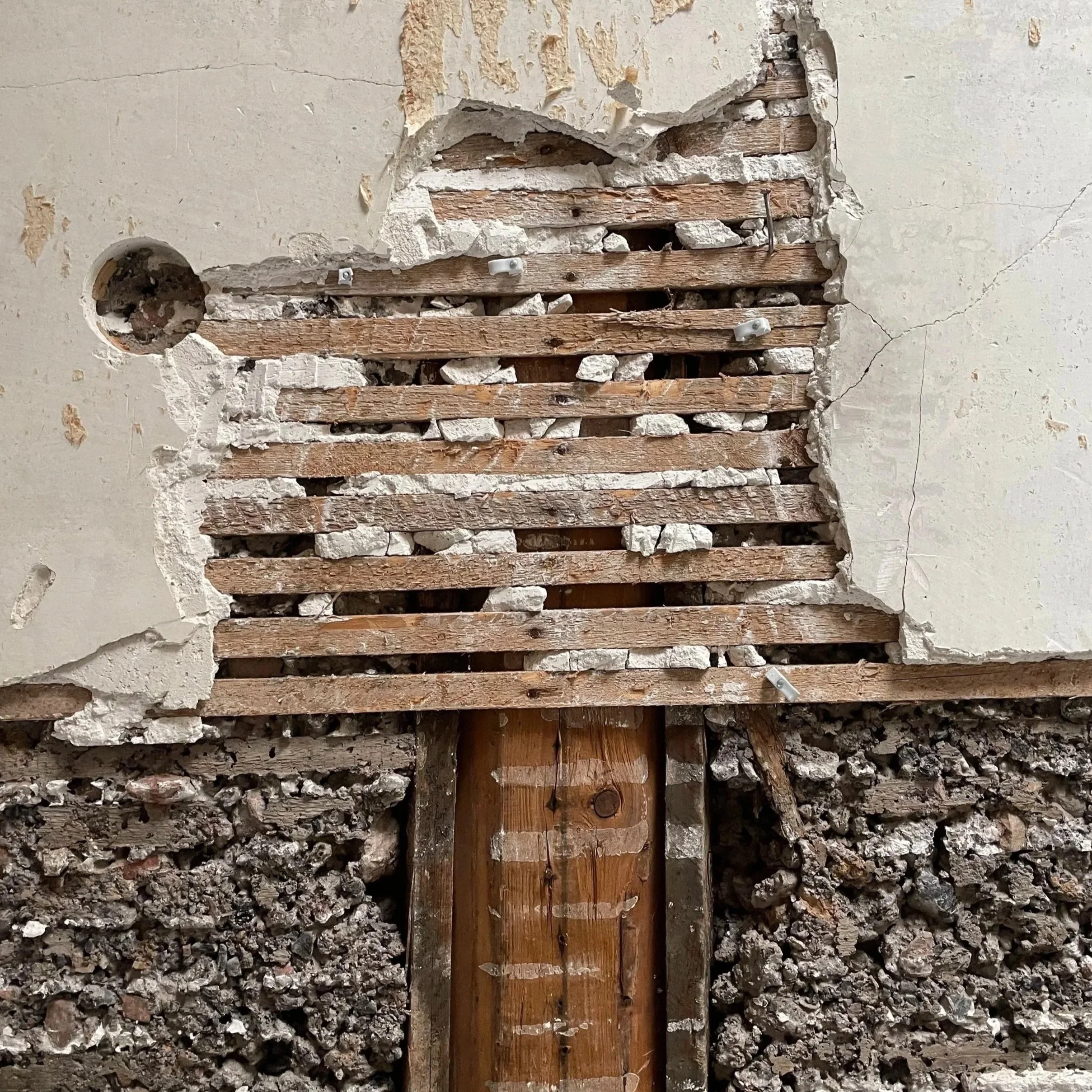 Ein Loch in einer verputzten Wand, das teilweise entfernt wurde, offenbart Holz- und Ziegelstrukturen im Inneren, mit Schrauben und Drähten. Der Bereich darunter ist mit kleinen Steinen und Erde gefüllt.
