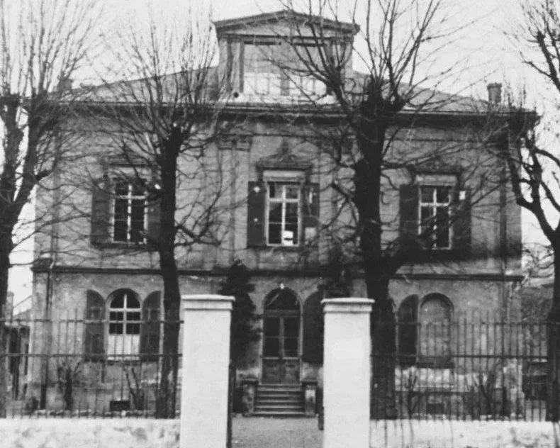 Ein altes, zweistöckiges Haus mit klassischer Architektur, umgeben von einem Zaun und Bäumen ohne Laub, wahrscheinlich im Winter.