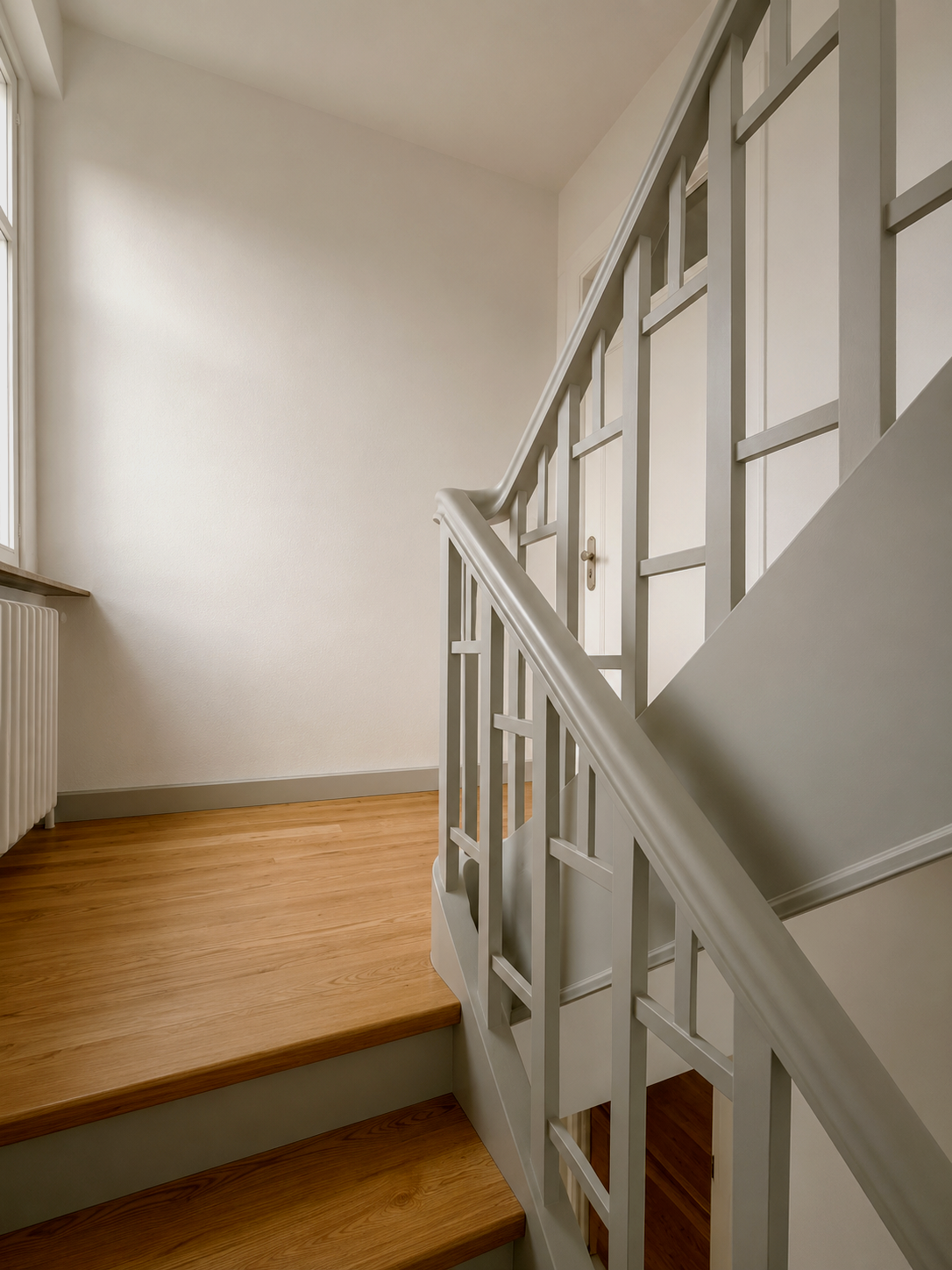 Innenaufnahme einer Treppe mit Holzstufen und weißen Geländer in einem Haus, neben einem Fenster und einem Heizkörper.