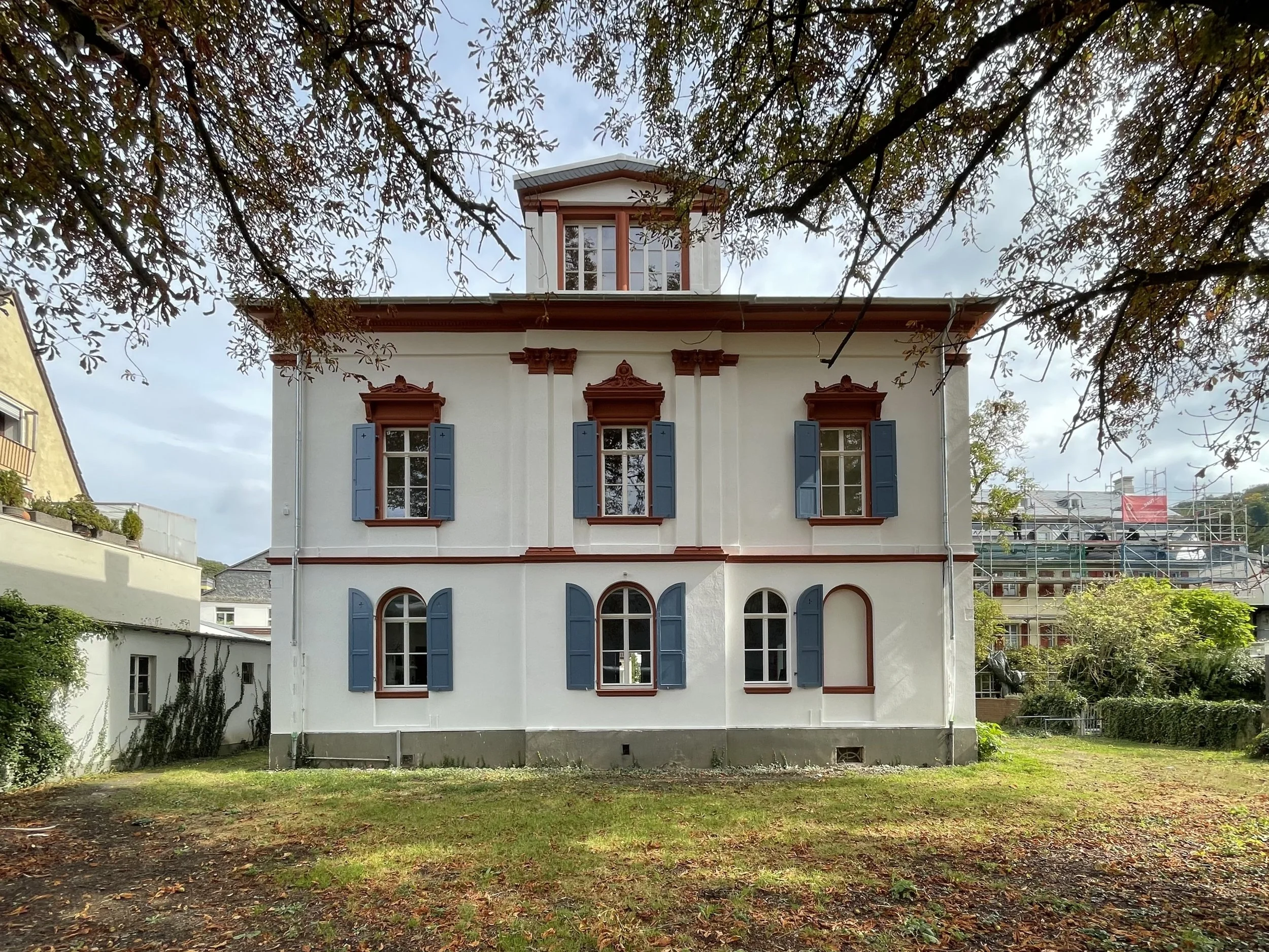 Ein mehrstöckiges Haus mit blauen Fensterläden, eingerahmt von Bäumen und einem Rasen im Vordergrund.