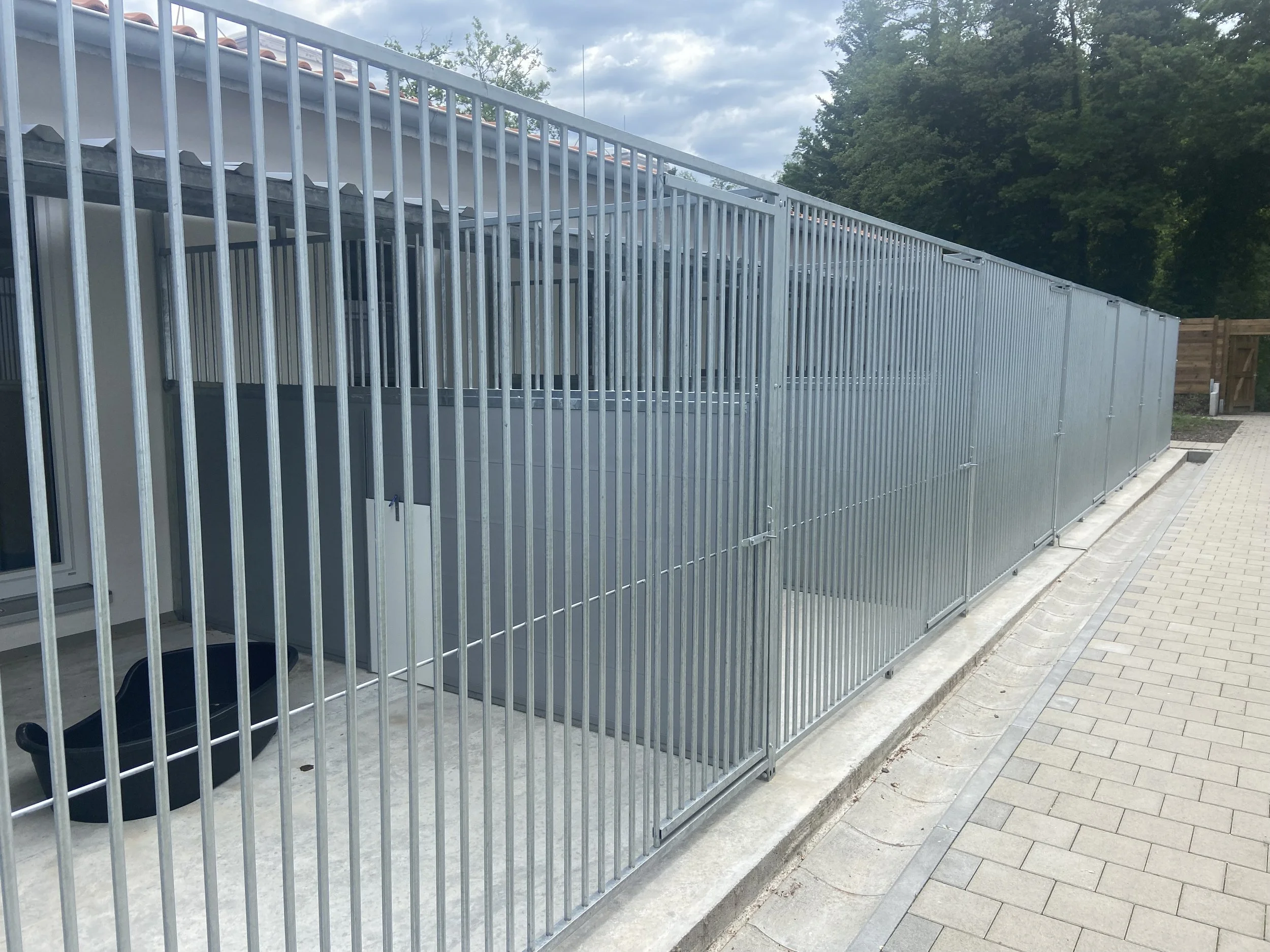 Ein Metallzaun, der eine Tierkiste umgibt, mit einem Hundekorb im Inneren, vor einem Gehweg und umgeben von Bäumen.