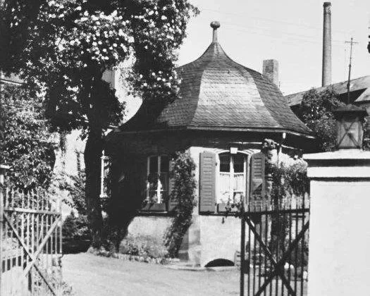 Schwarz-weises Foto eines kleinen Hauses mit einer runden, turmähnlichen Dachform, umgeben von Zäunen und Bäumen.