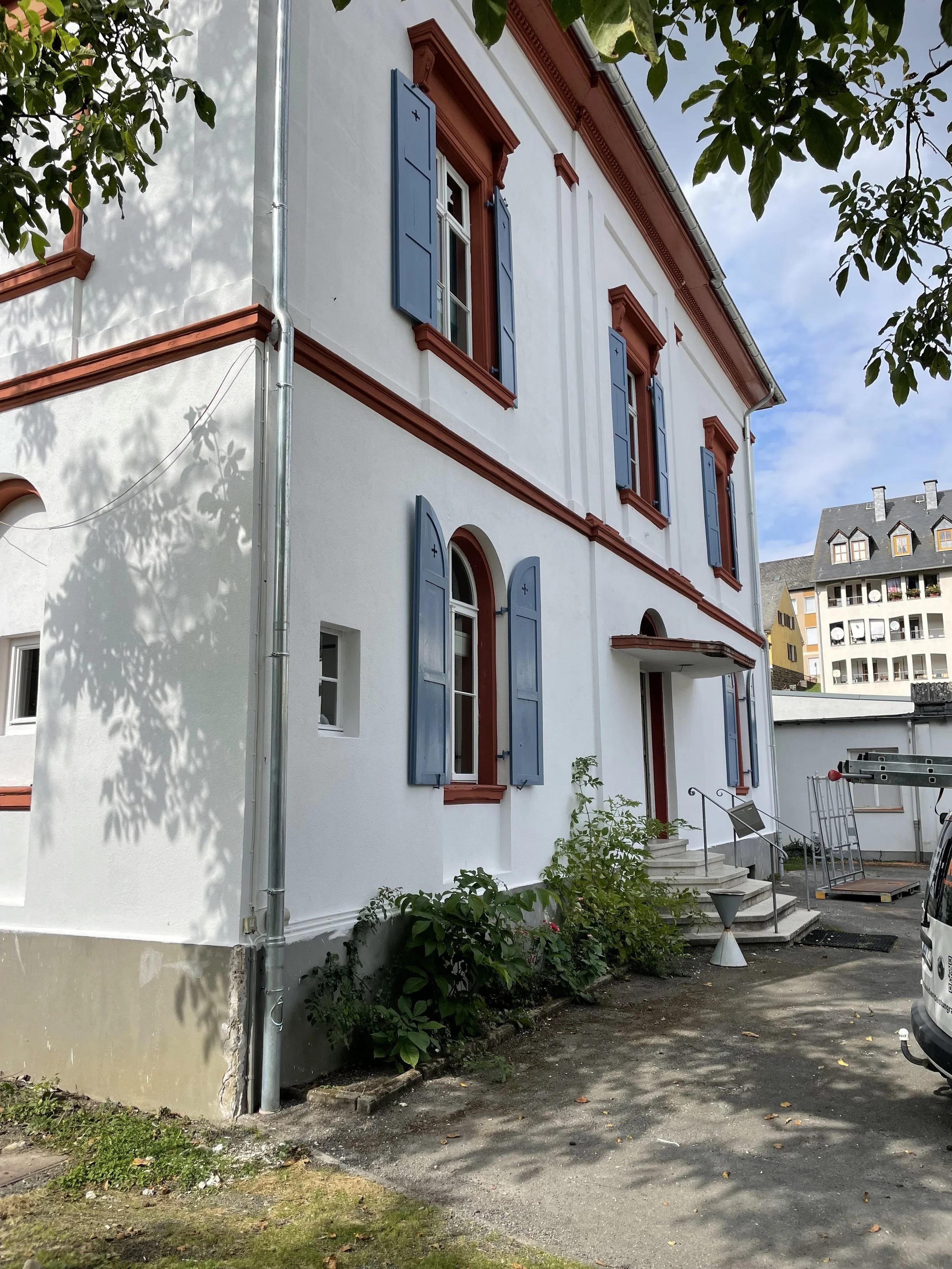 Ein weißes Haus mit roten und blauen Fensterläden, einer Treppe vor dem Eingang, und einem Überblick über die Umgebung mit weiteren Häusern und Bäumen bei sonnigem Himmel.