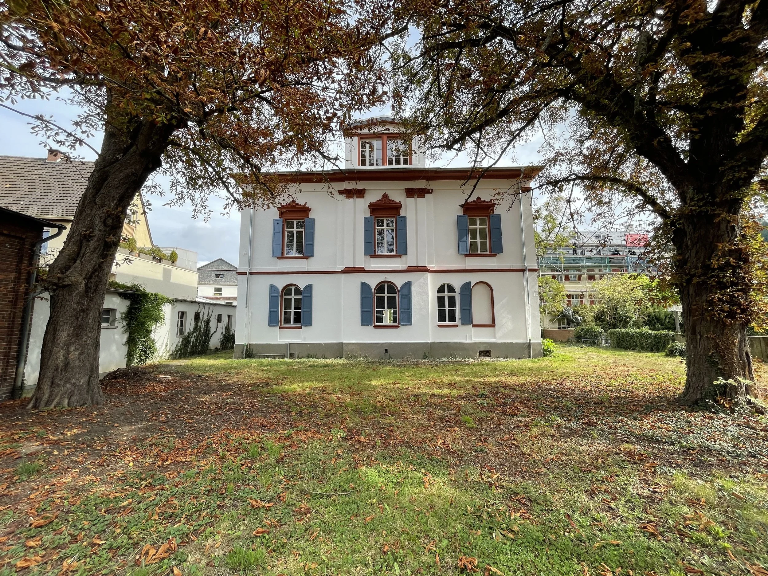 Ein Haus mit weißen Wänden, blauen Fensterläden und einem rotbraunen Dach, umgeben von Bäumen mit braunen und grünen Blättern, im Vordergrund ein Rasen und herbstlich gefärbte Blätter.