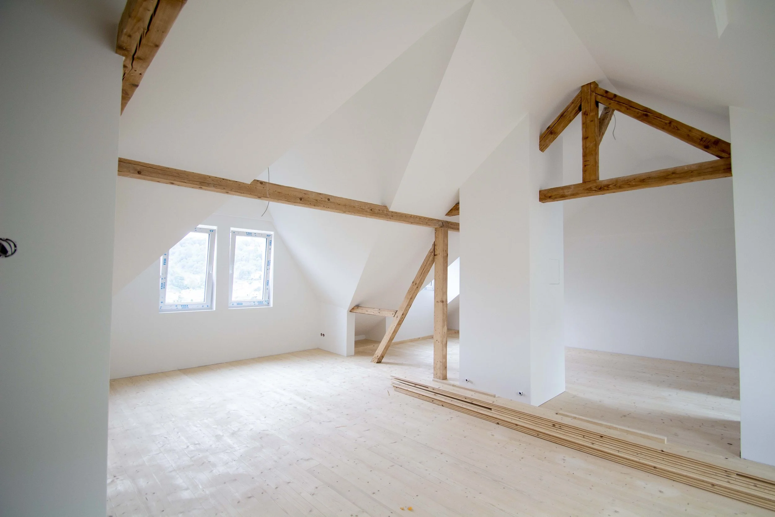 Ein leerer Raum mit weißen Wänden und großen Fenstern. Es gibt sichtbare Holzbalken und ein Holzpodest im Raum.