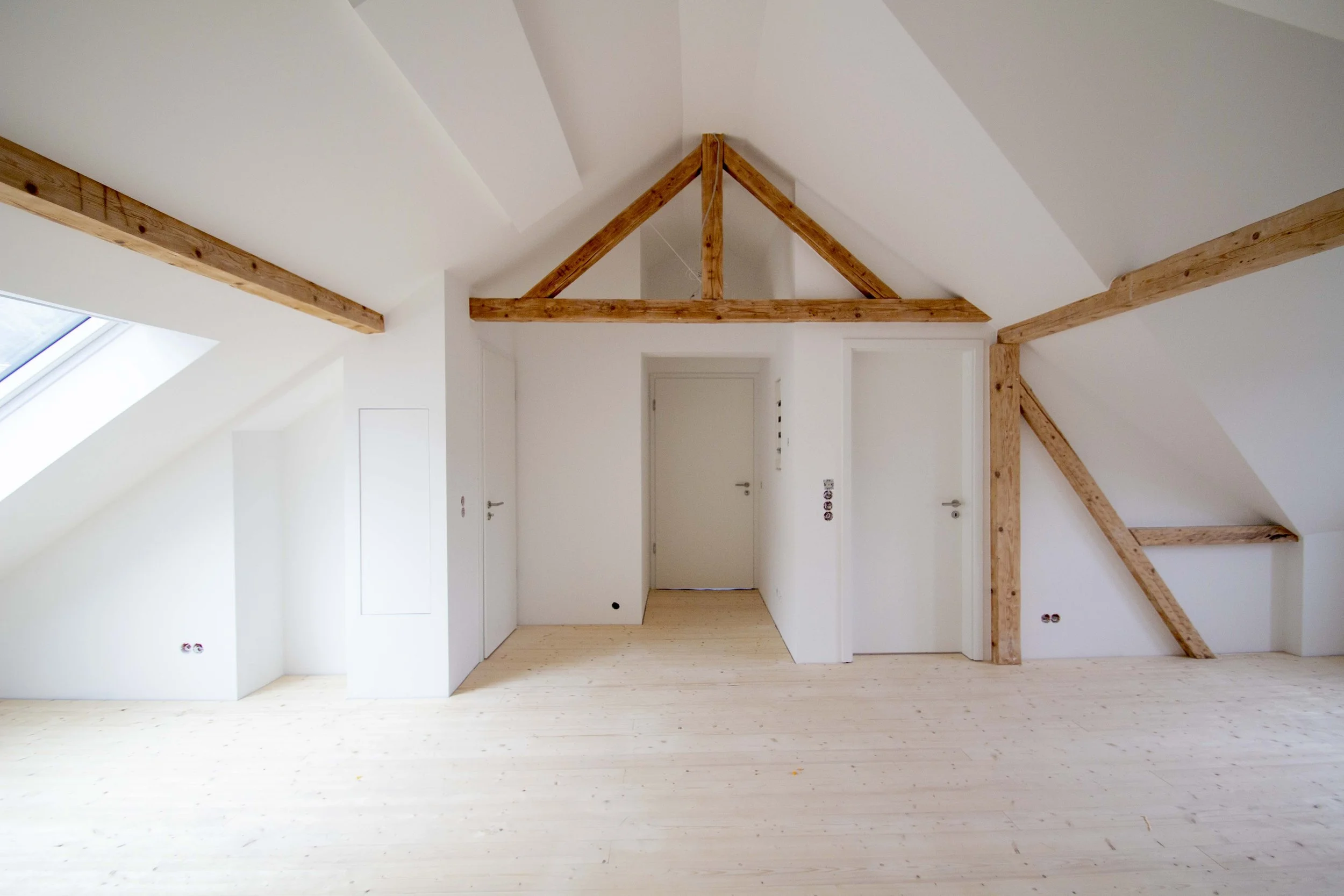 Ein leeres, modern eingerichtetes Dachzimmer mit weißen Wänden, Holzbalken an der Decke und einem Holzboden.