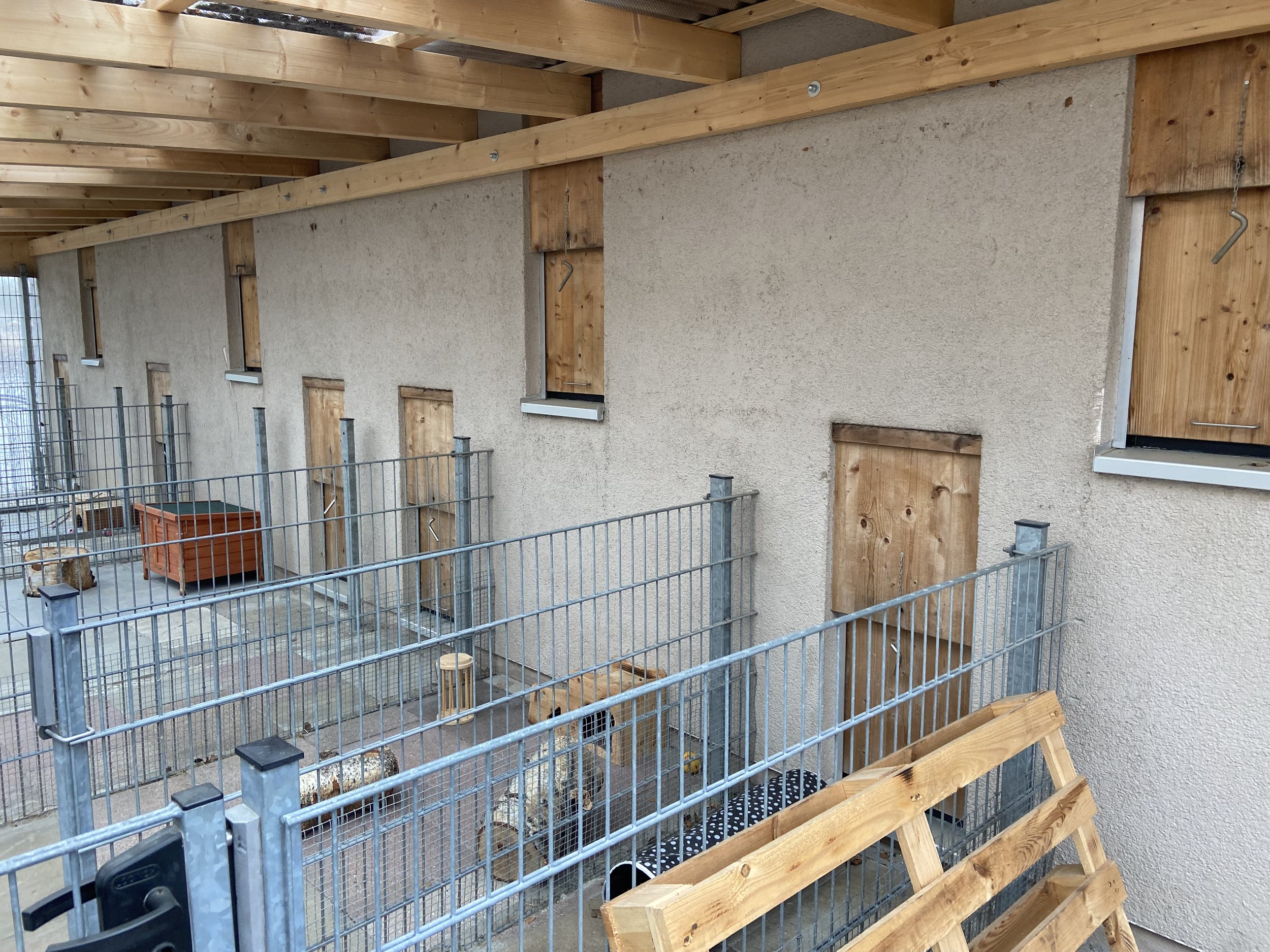 Baustelle mit Wand aus Holz und Putz, Gitterzaun, Fensteröffnungen, Bauwerk im Rohbau, Bauholz und Baumaterialien sichtbar.