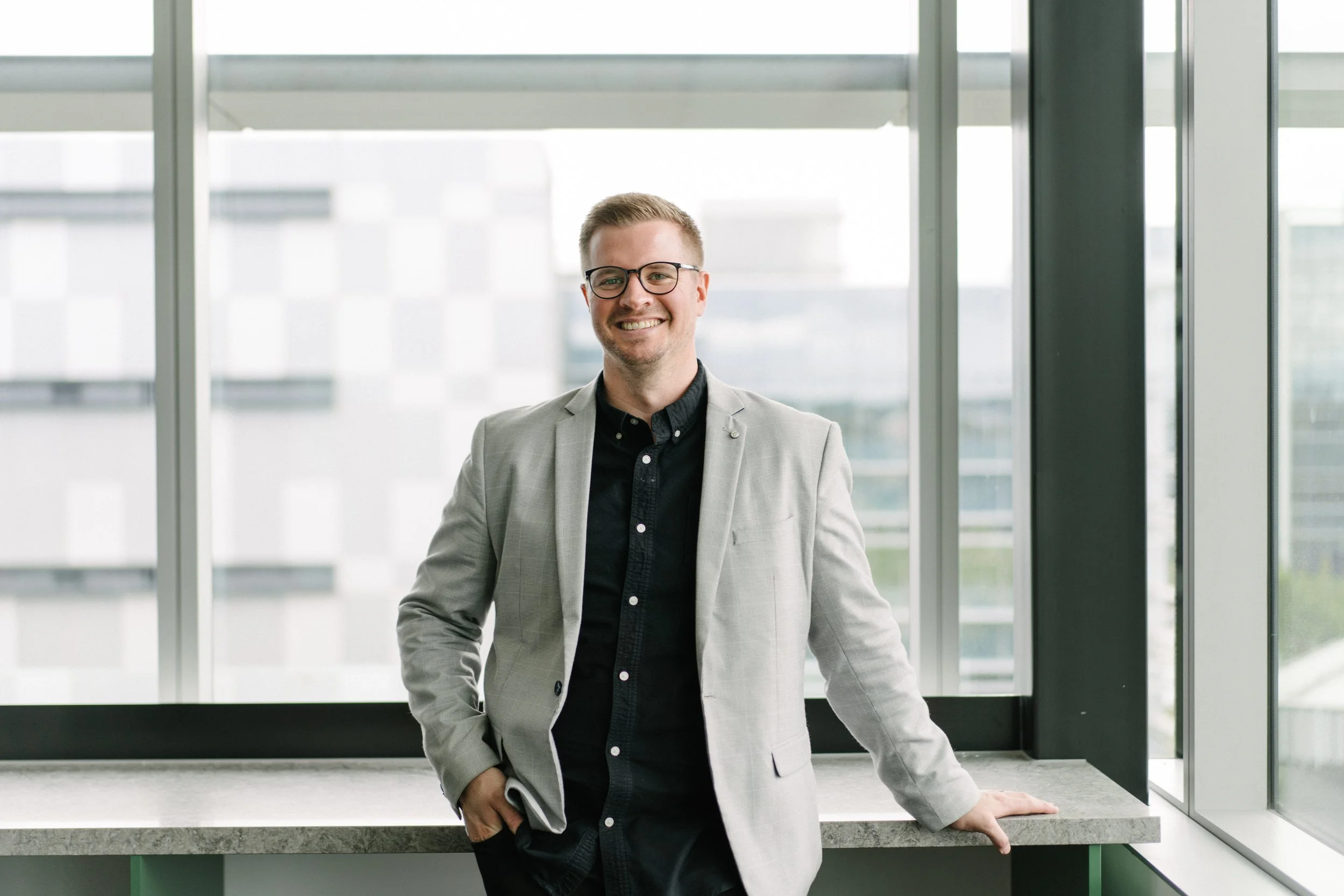 A young man with glasses, wearing a light gray blazer and black shirt, smiling and standing by a window with tall buildings outside.