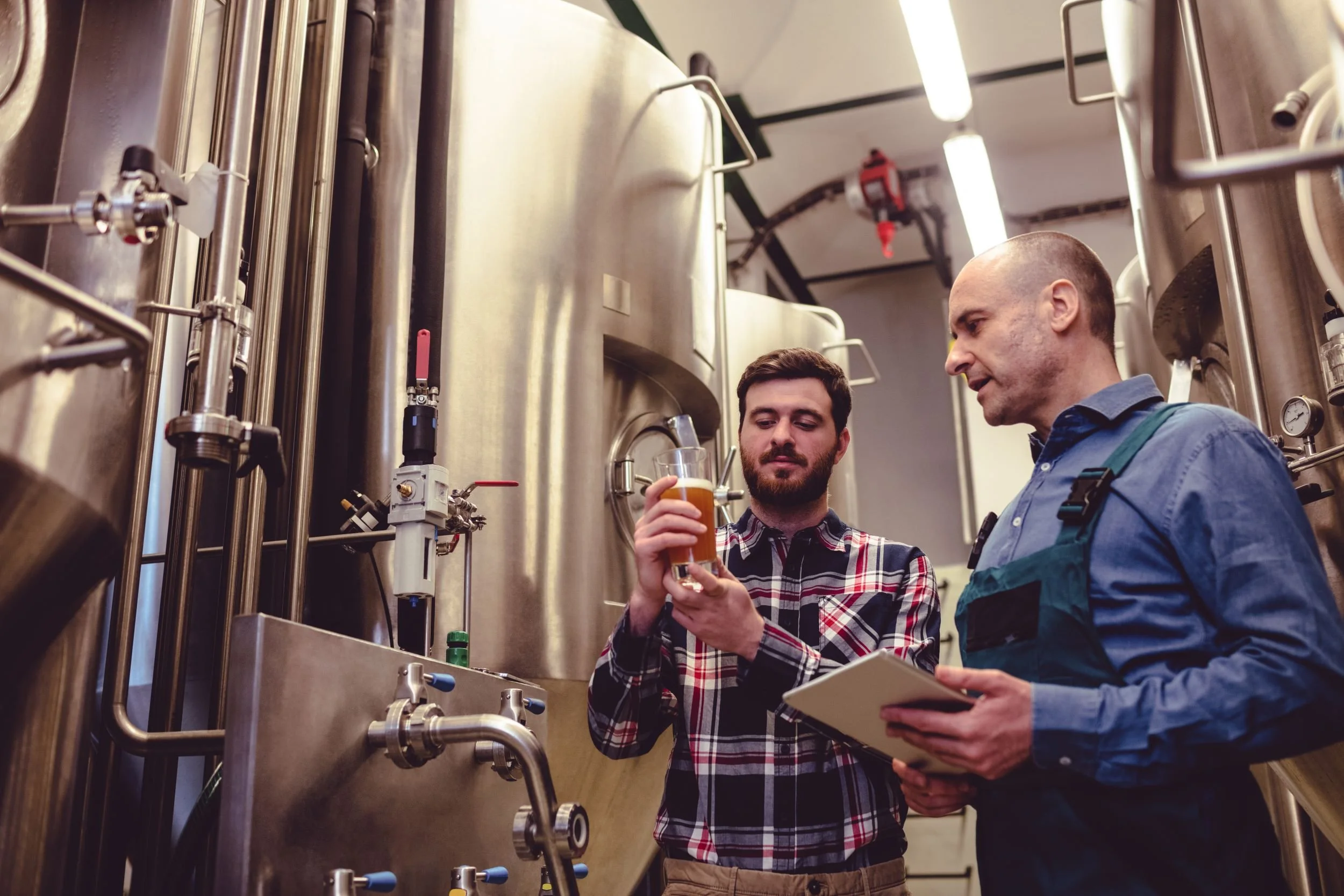 Zwei Brauer kontrollieren ein Glas Bier vor großen Edelstahl-Gärtanks in einer Brauerei