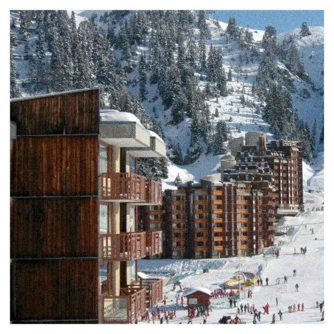 immeubles en bois et en béton au bord d'une station de ski, avec des skieurs et snowboarders dans la neige, entourés de montagnes enneigées et de sapins