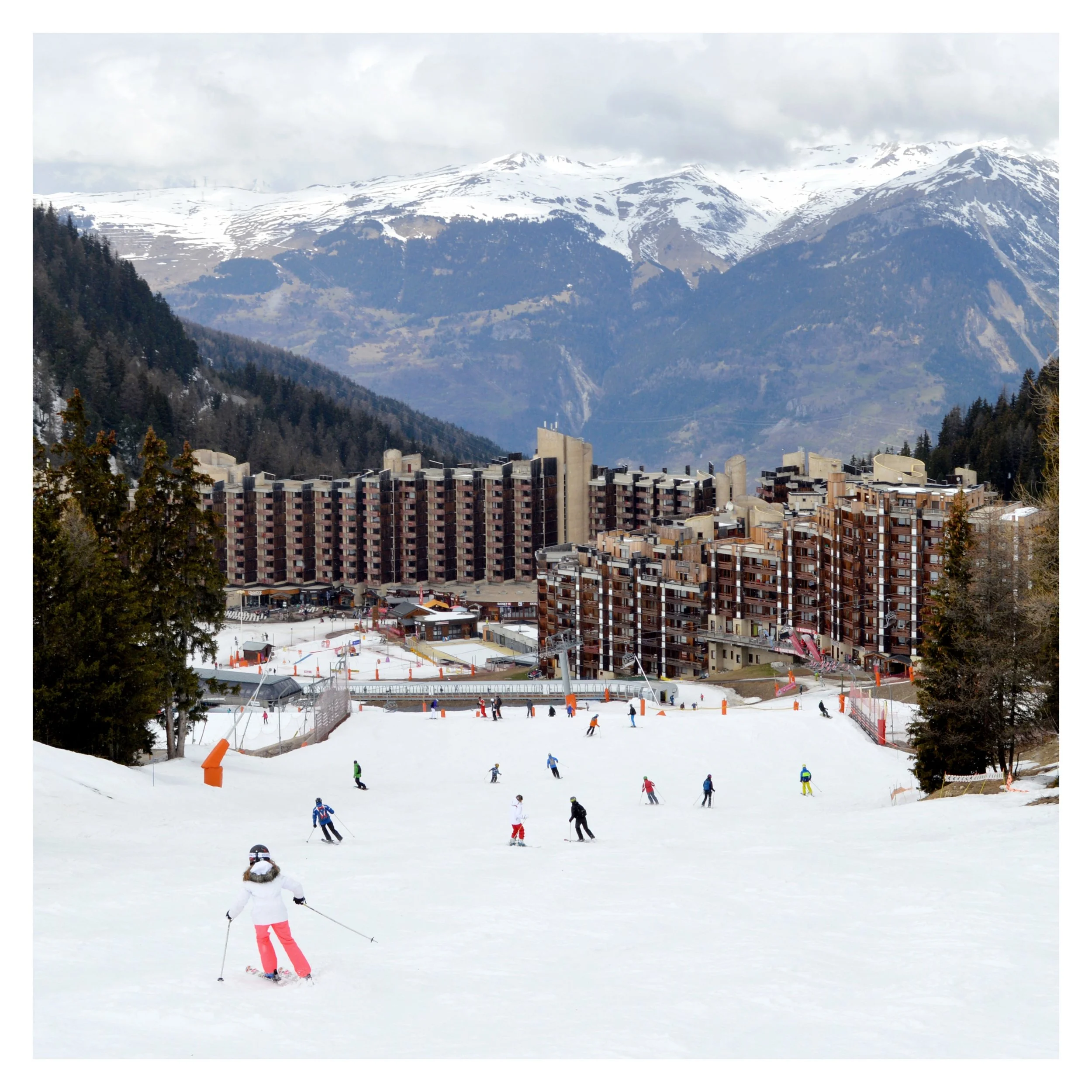 Des skieurs en train de descendre une piste de ski dans une station de montagne enneigée avec des bâtiments modernes en arrière-plan et des montagnes enneigées sous un ciel nuageux.