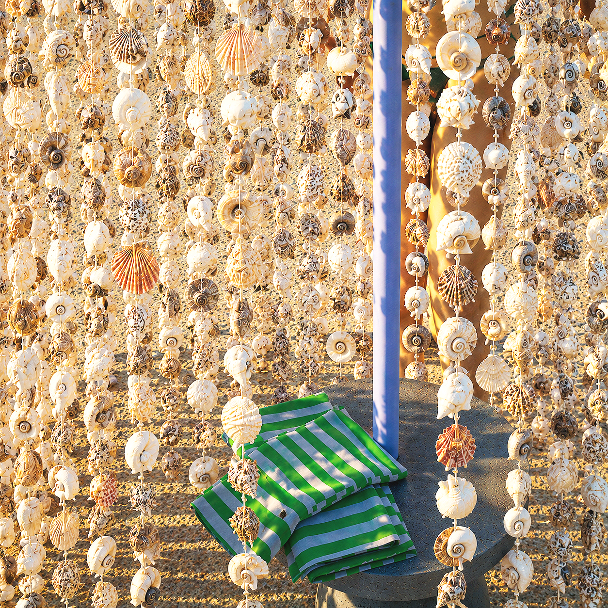 Des guirlandes de coquillages suspendues en éventail dans un décor de plage, avec un parasol en paille orange et des matelas ou coussins à rayures vertes et blanches posés sur le sable.