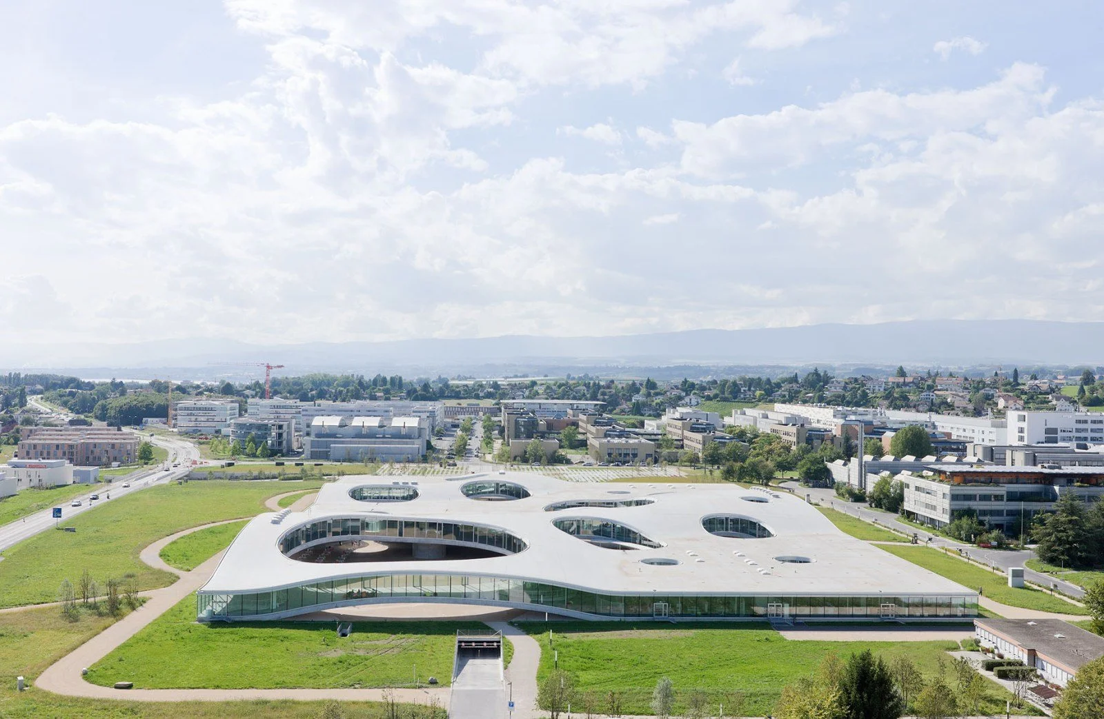 Photo d'un bâtiment futuriste avec une architecture innovante, caractérisée par une toiture ondulée avec plusieurs oculus, situé dans un environnement urbain avec des immeubles résidentiels et commerciaux en arrière-plan, sous un ciel partiellement n