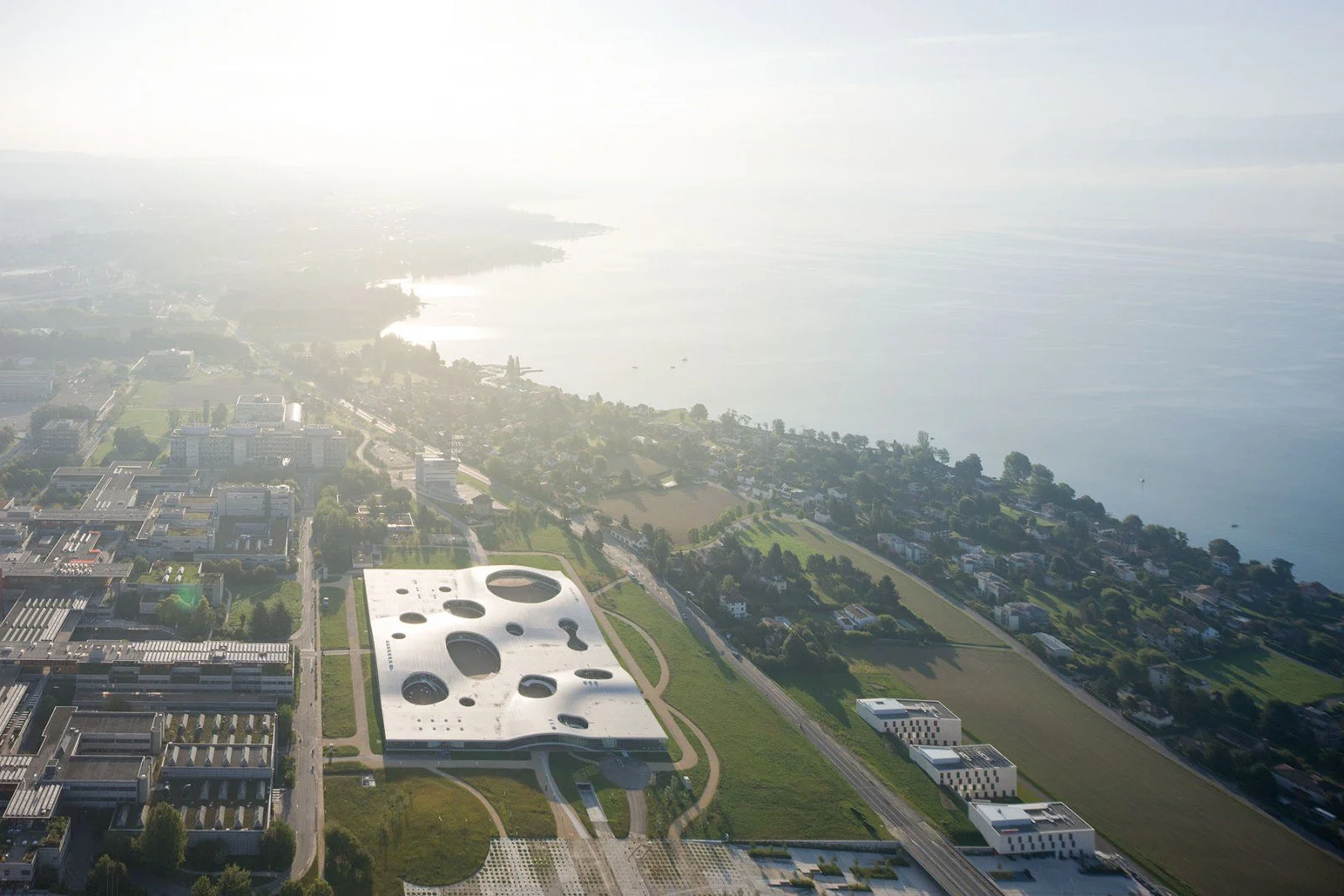 Vue aérienne d'une ville avec un lac à l'horizon, un bâtiment moderne avec des formes ovales, routes et zones vertes.