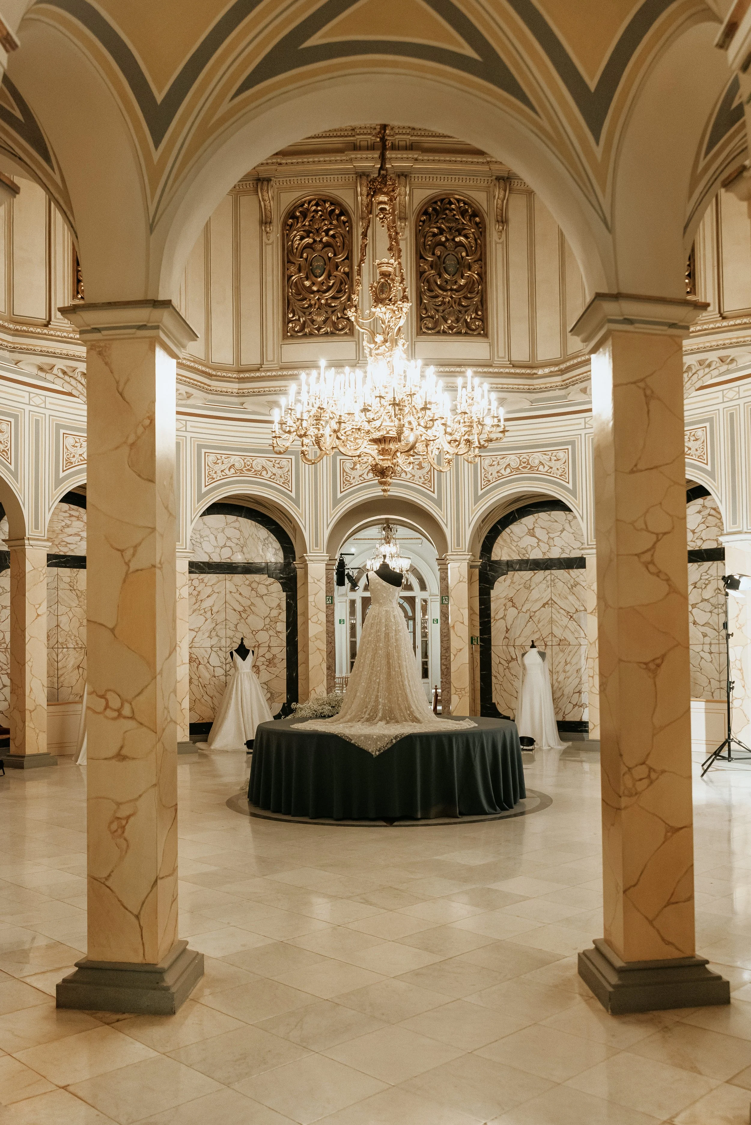 Vestido de novia en exhibición en una sala elegante y lujosa con columnas de mármol y candelabro grande en el techo, en un evento de moda o tienda de novias.