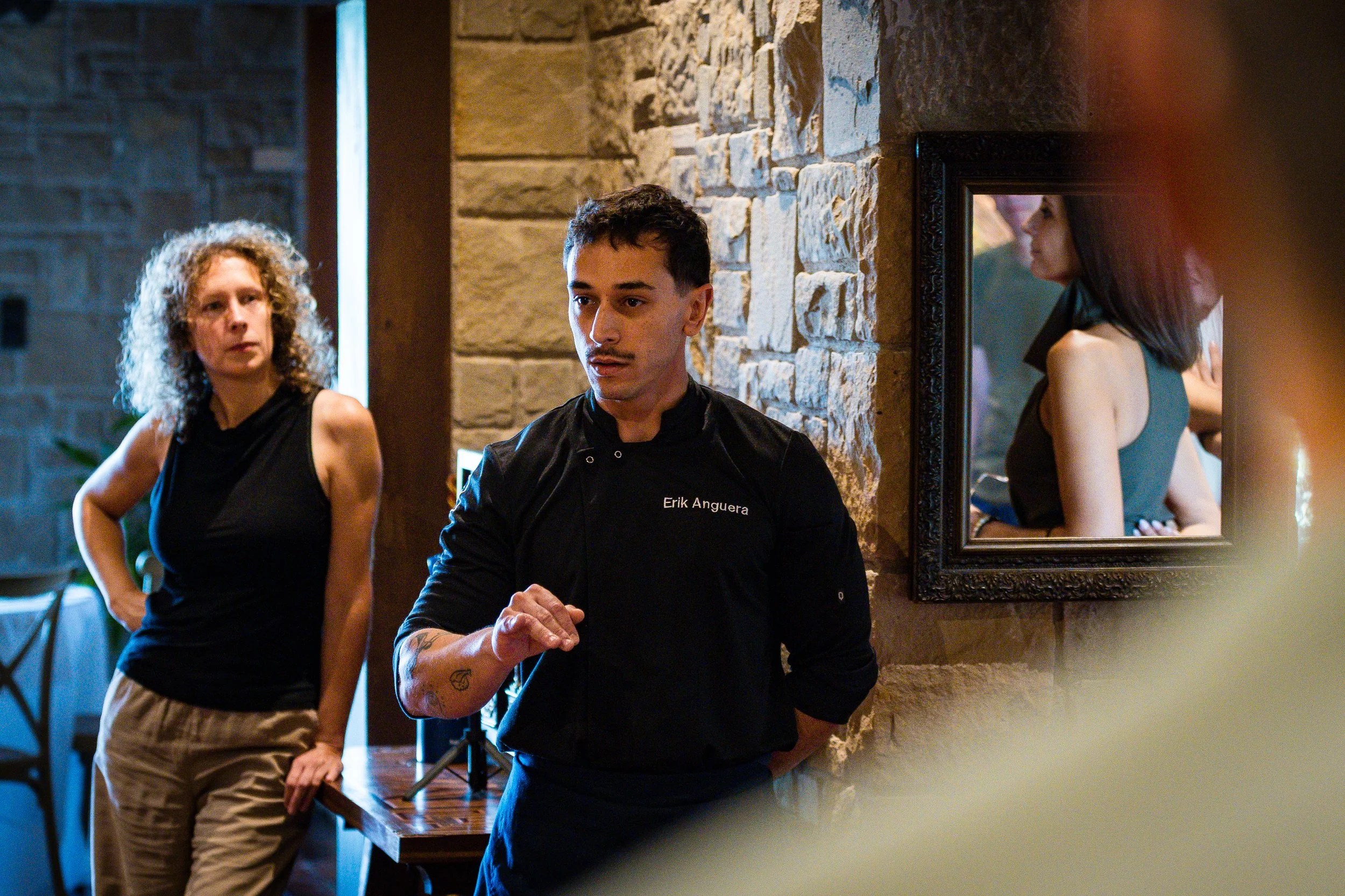 Joven hombre en uniforme de chef hablando en un restaurante, con dos mujeres en el fondo y un espejo que refleja a una mujer.