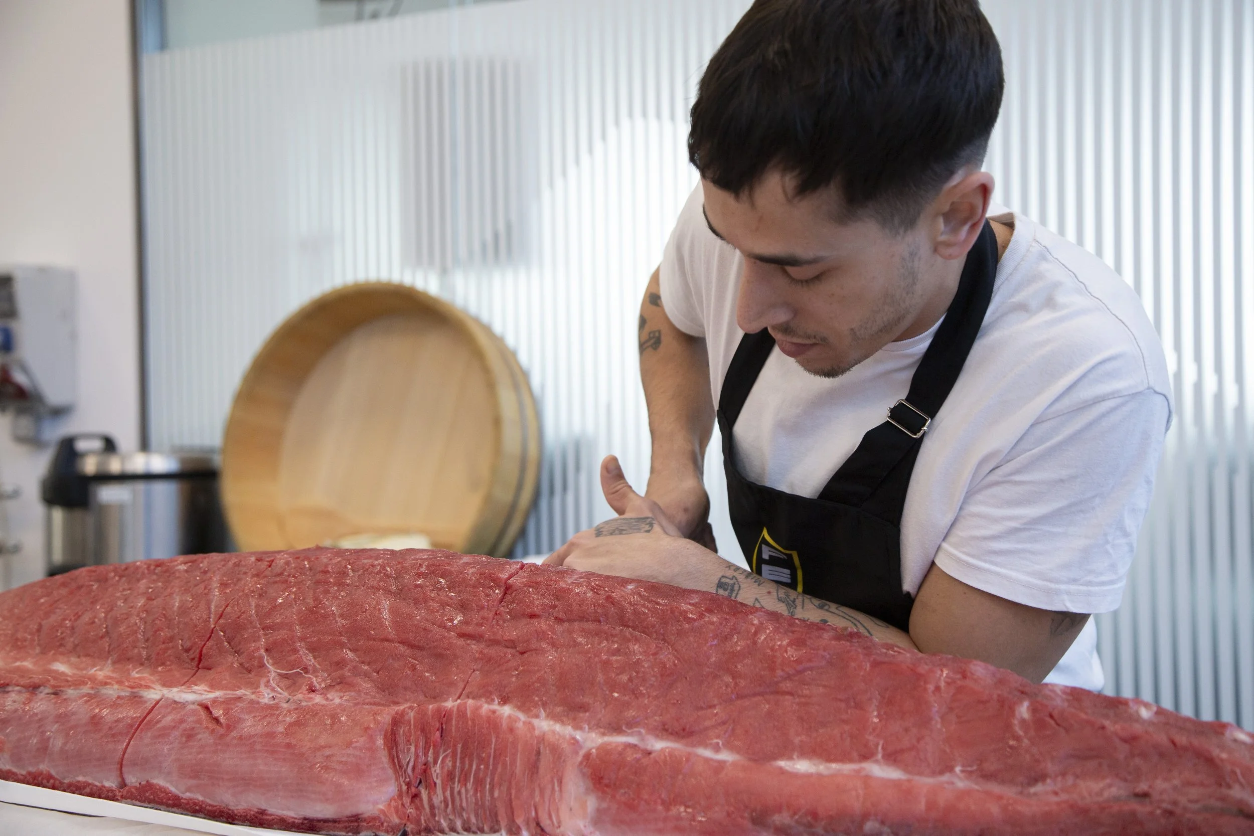 Hombre cortando carne en una cocina. En primer plano, un trozo grande de carne y en el fondo, el hombre con camiseta blanca y delgada, con cabello oscuro y corto, trabajando en la preparación de la carne.