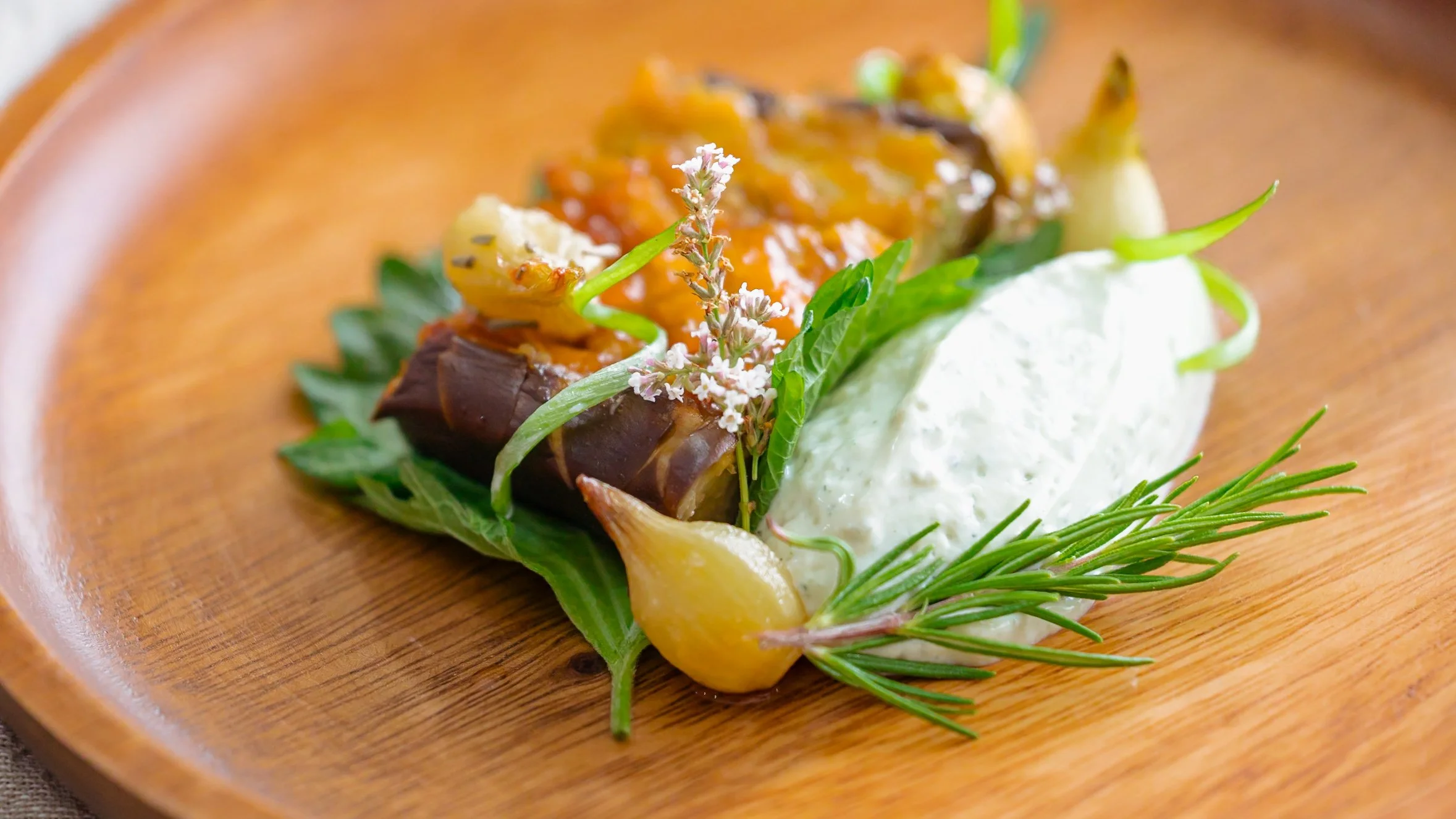 Plato vegetariano con berenjena asada al miso, crema de tofu, vegetales verdes, y decorado con flores y hierbas aromáticas en un plato de madera.