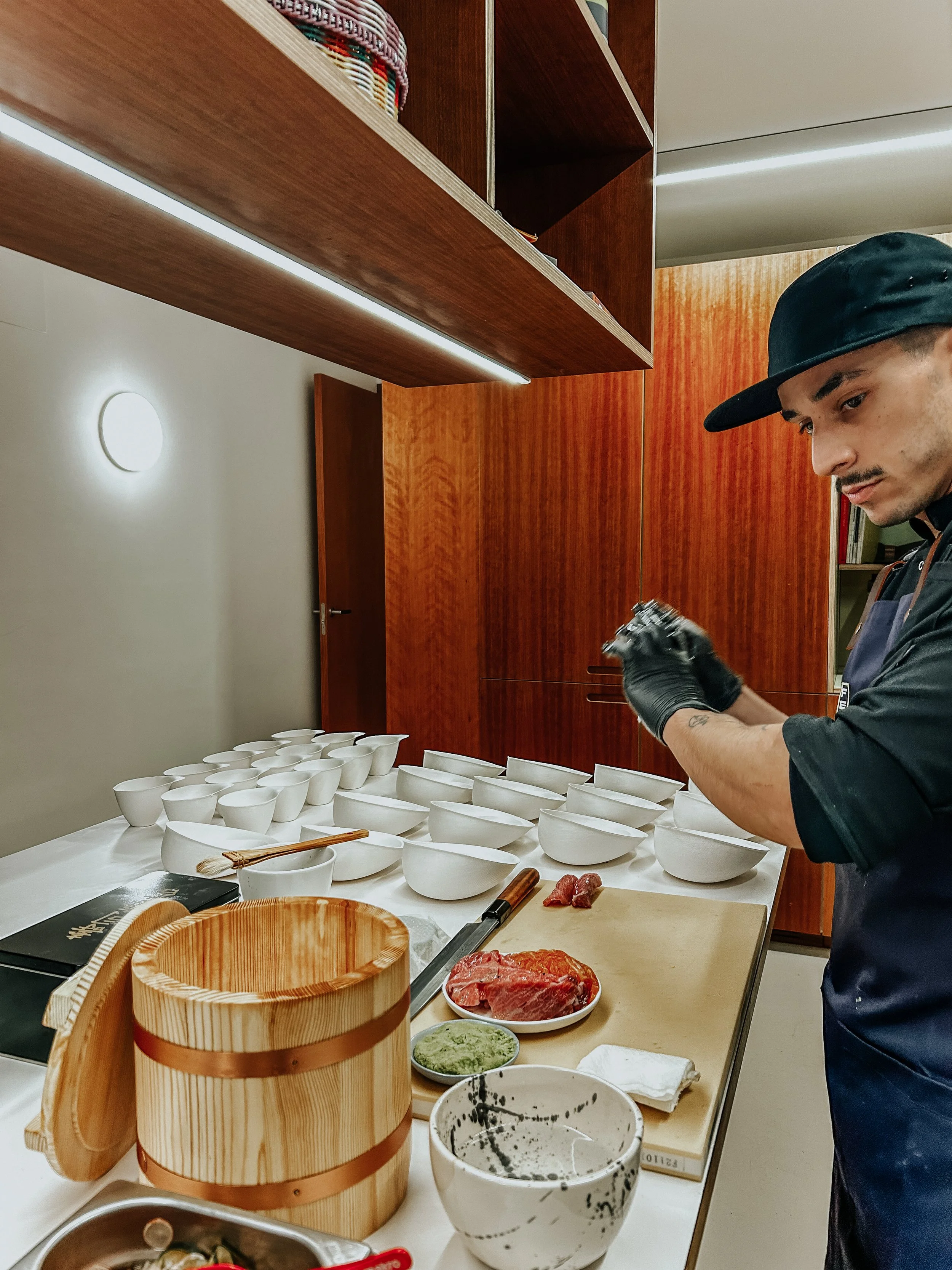 Un hombre preparando sushi en una cocina moderna con platos blancos alineados en la mesa, ingredientes como carne, wasabi, y utensilios japoneses visibles.