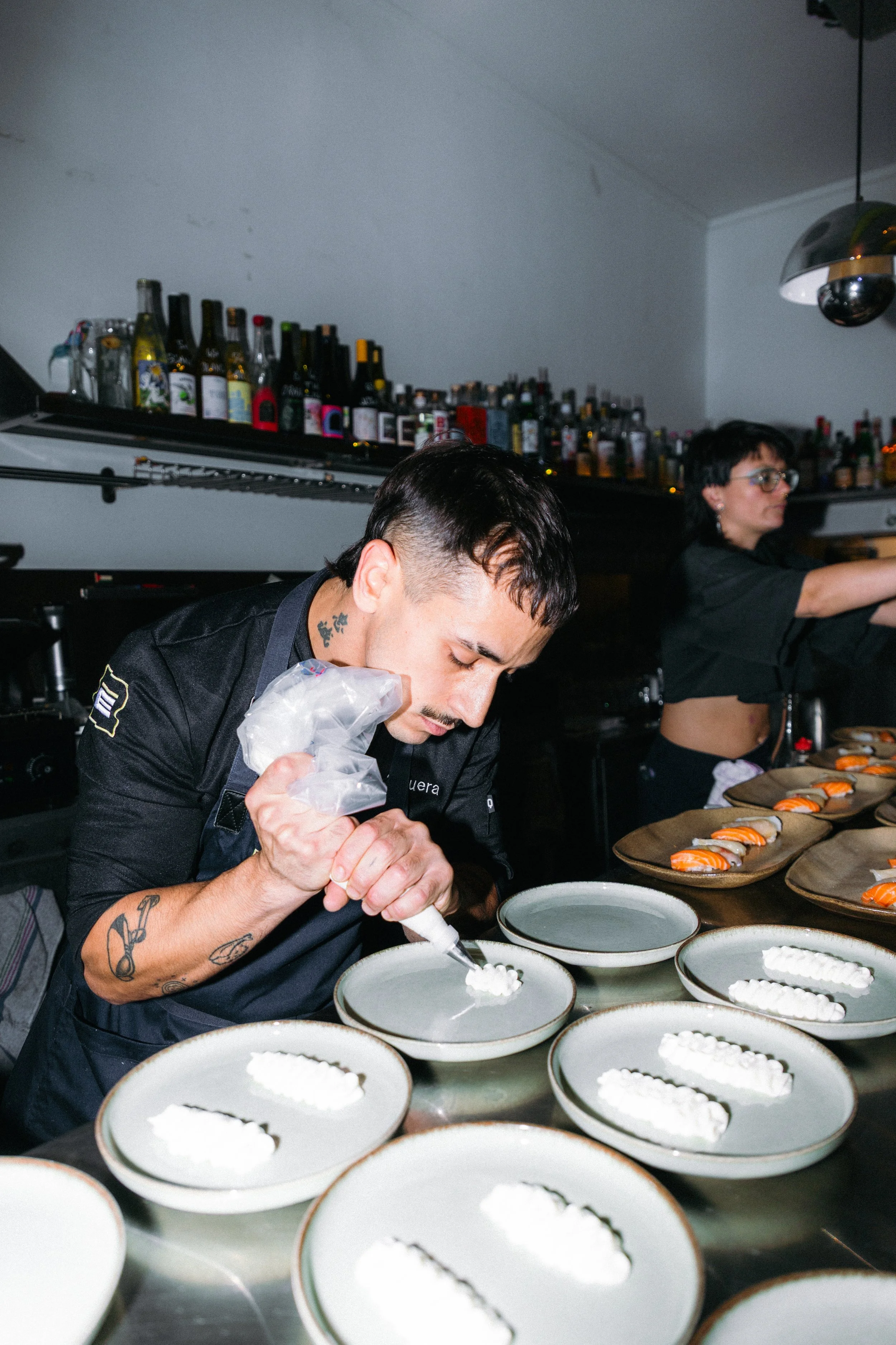 Un chef decorando platos de sushi en una cocina profesional, con botellas de bebidas en una estantería al fondo.