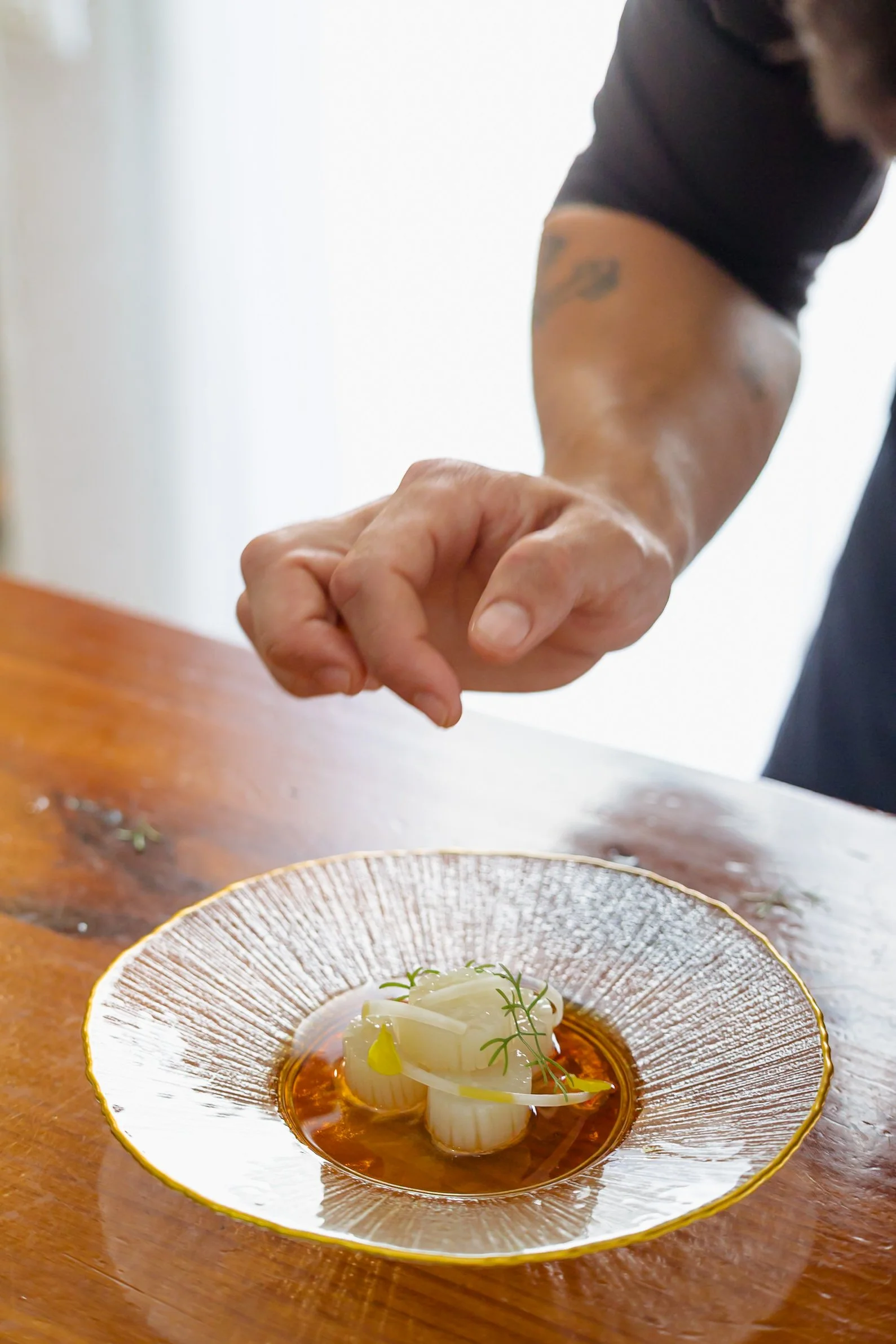 Primer plano de un plato de comida elegante con un caldo oscuro, acompañado de una porción de cebolla en rodajas y unas ramitas de eneldo, en un plato decorativo con borde dorado sobre una mesa de madera.