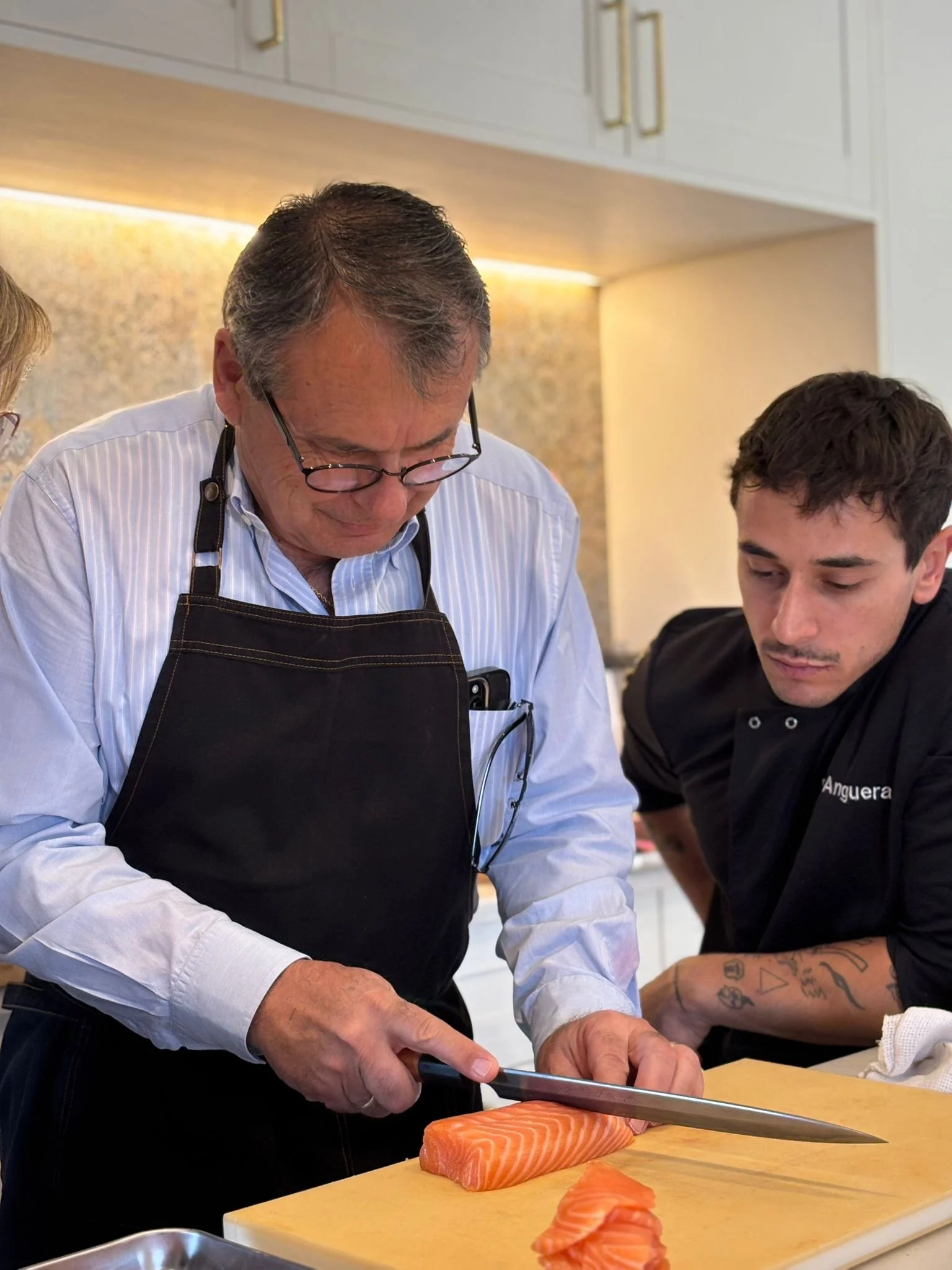 Dos personas, un hombre mayor con gafas y cabello gris y un joven con tatuajes en el brazo, están cortando un filete de salmón en una cocina moderna.
