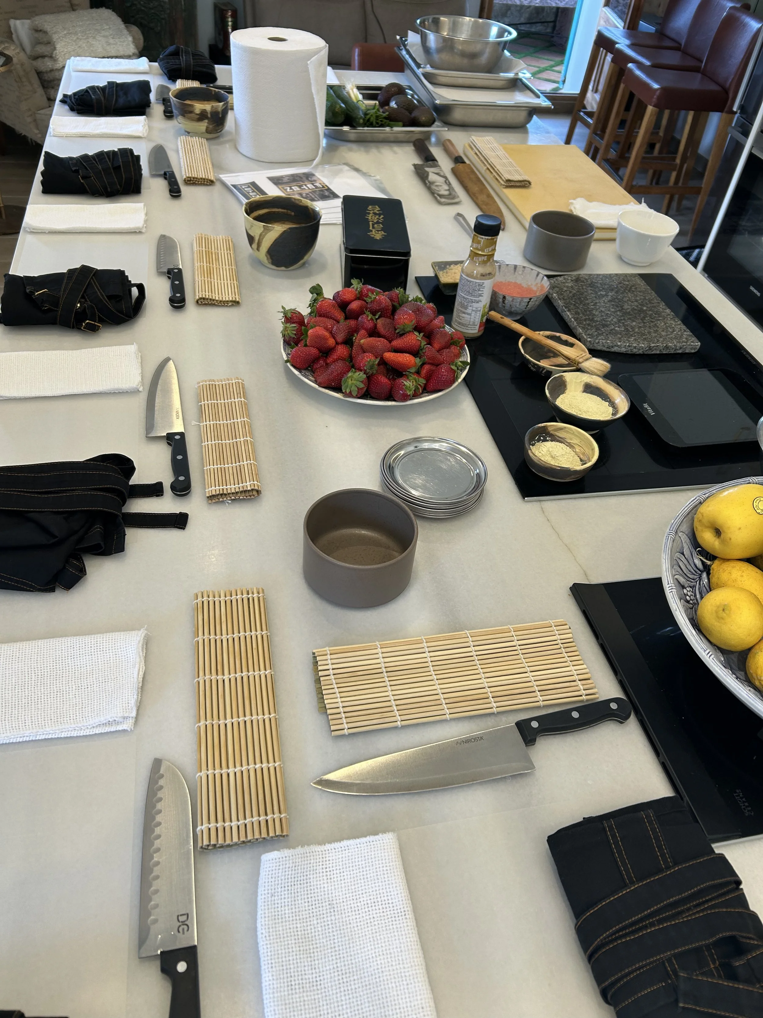 Mesa de cocina preparada para sushi con ingredientes y utensilios, incluyendo fresas, cuchillos, palillos, tazas, y otros utensilios de cocina.