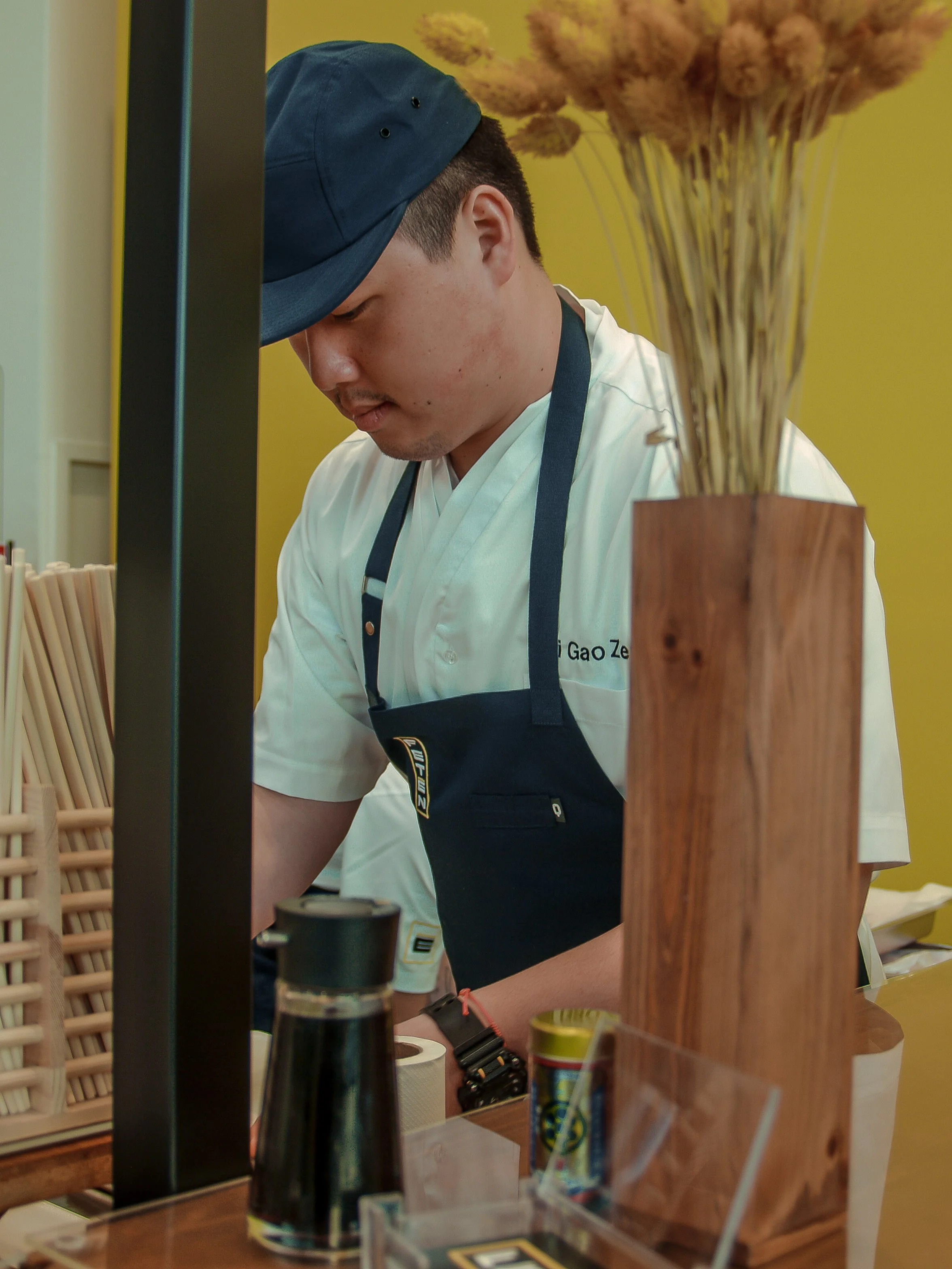Joven cocinero de espaldas, con gorra azul y delantal, en un restaurante, frente al mostrador con utensilios y productos de cocina.