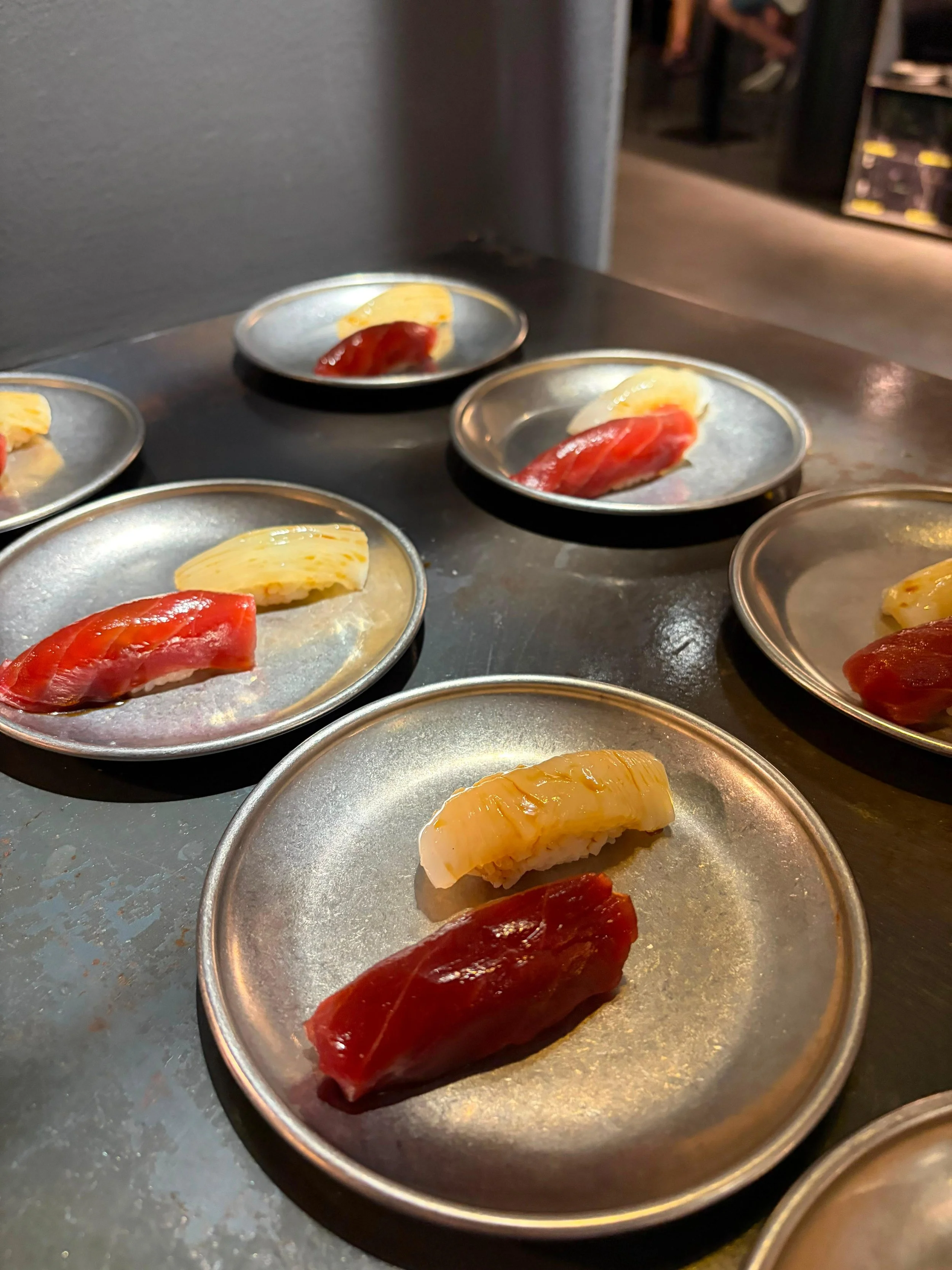 Variedad de piezas de sushi sobre platos de metal en una barra de sushi.