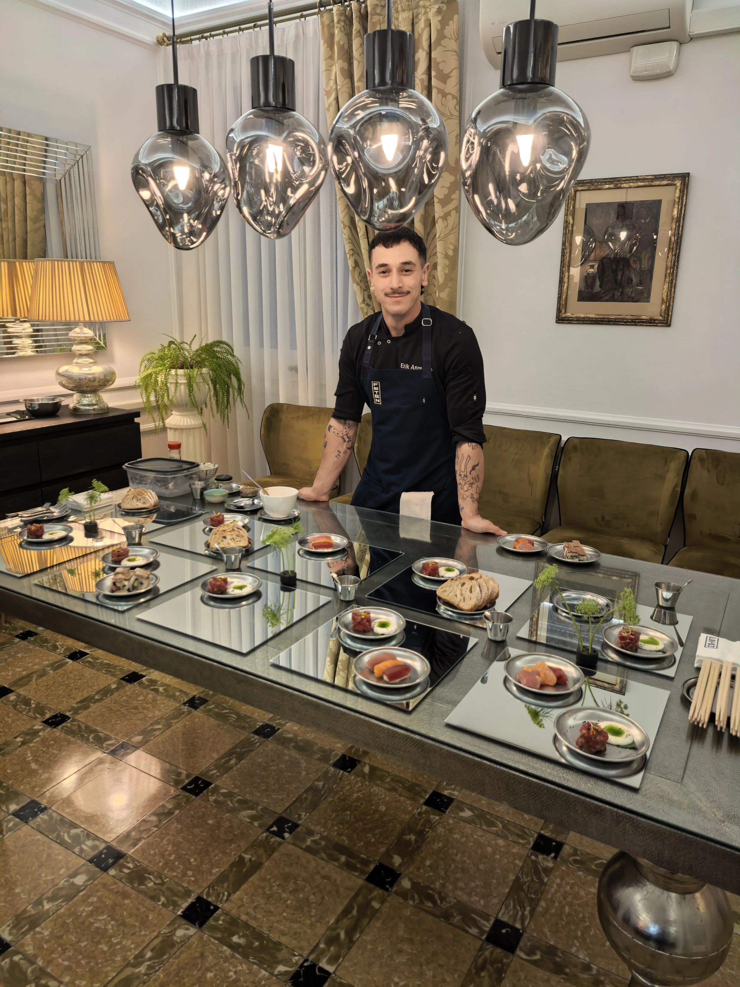 Hombre con barba y tatuajes en brazos, vistiendo un delantal azul oscuro, se encuentra detrás de una mesa de banquete con platos pequeños de comida, en un comedor elegante con luces modernas y decoraciones sofisticadas.