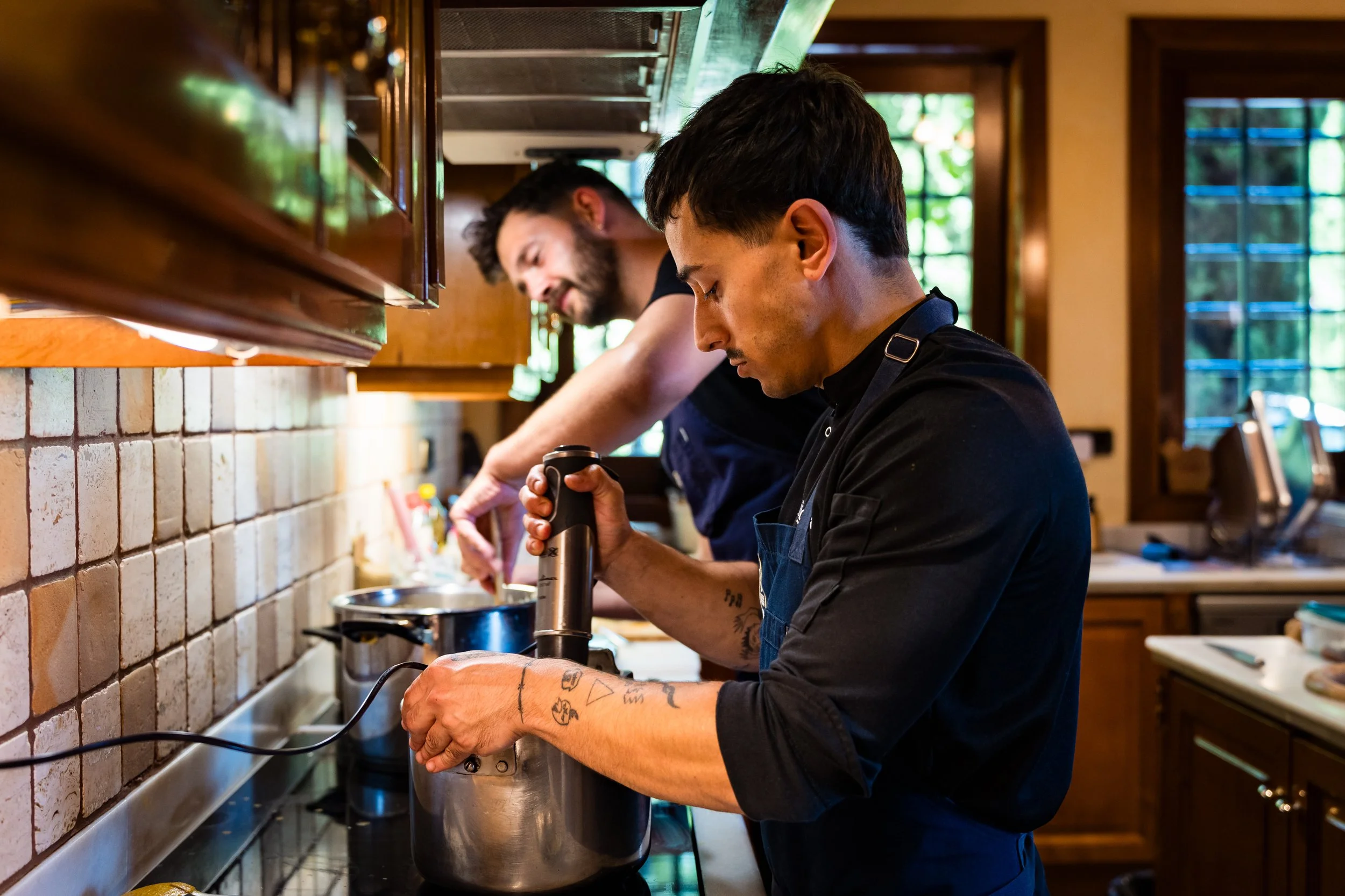 Dos hombres cocinando en una cocina de madera, uno de ellos sosteniendo una licuadora y el otro usando una cuchara grande en una olla.