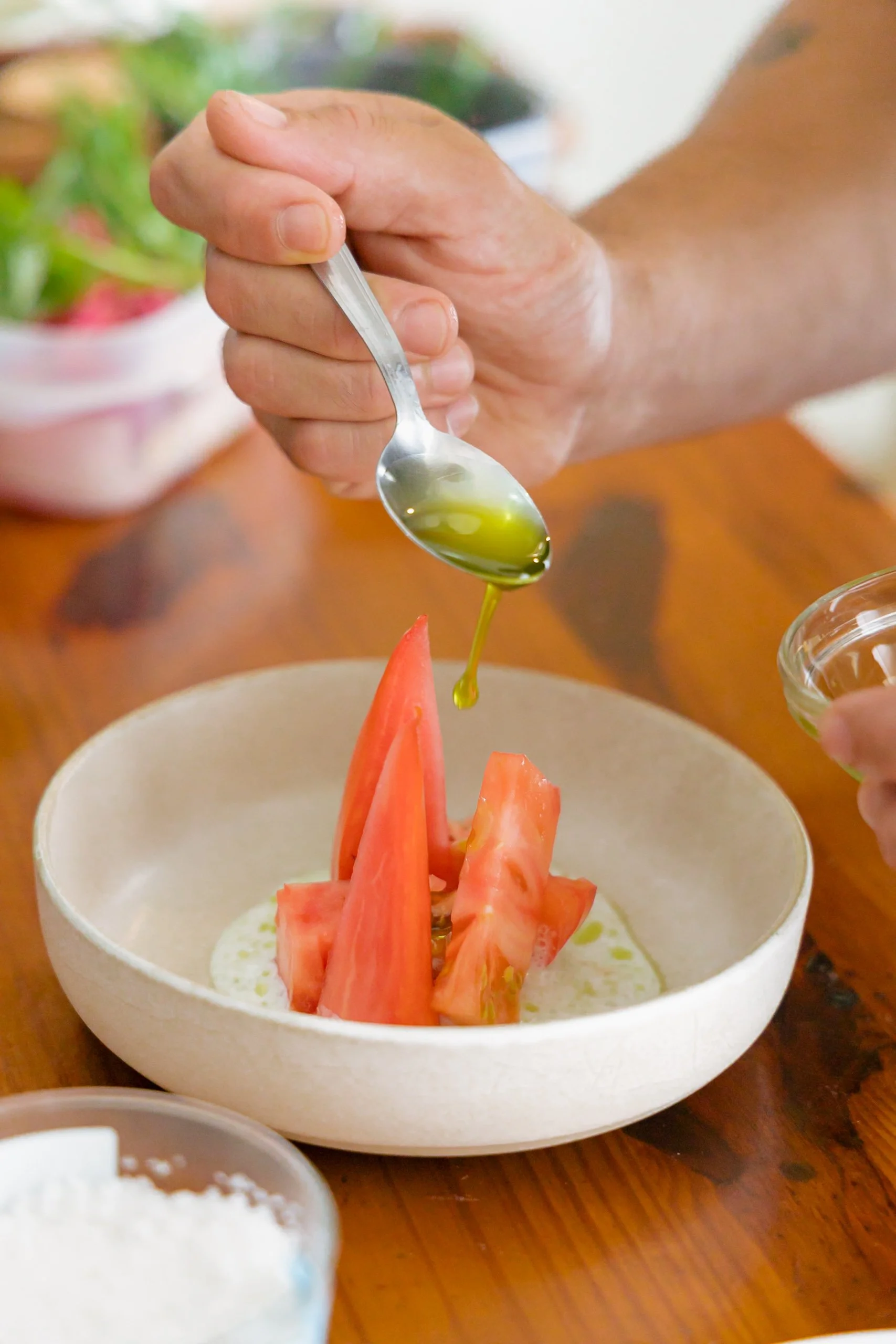 Una mano con un tenedor vertiendo aceite de oliva sobre una ensalada que tiene sandía y una salsa blanca en un recipiente de cerámica