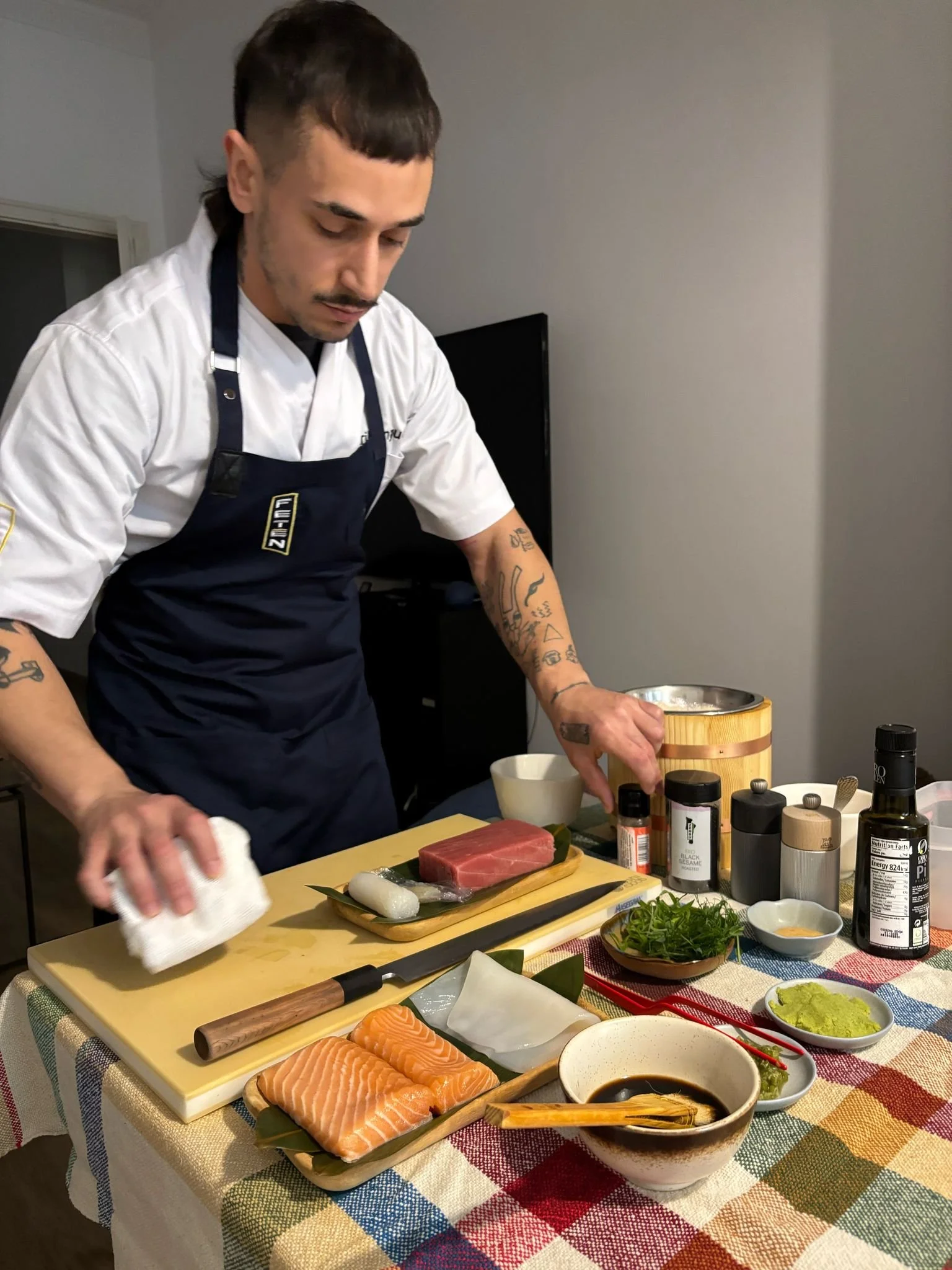 Un hombre preparando sushi en una mesa con ingredientes de pescado, el hombre lleva un delantal oscuro y una camiseta blanca, en un ambiente interno. Servicio omakase a domicilio.