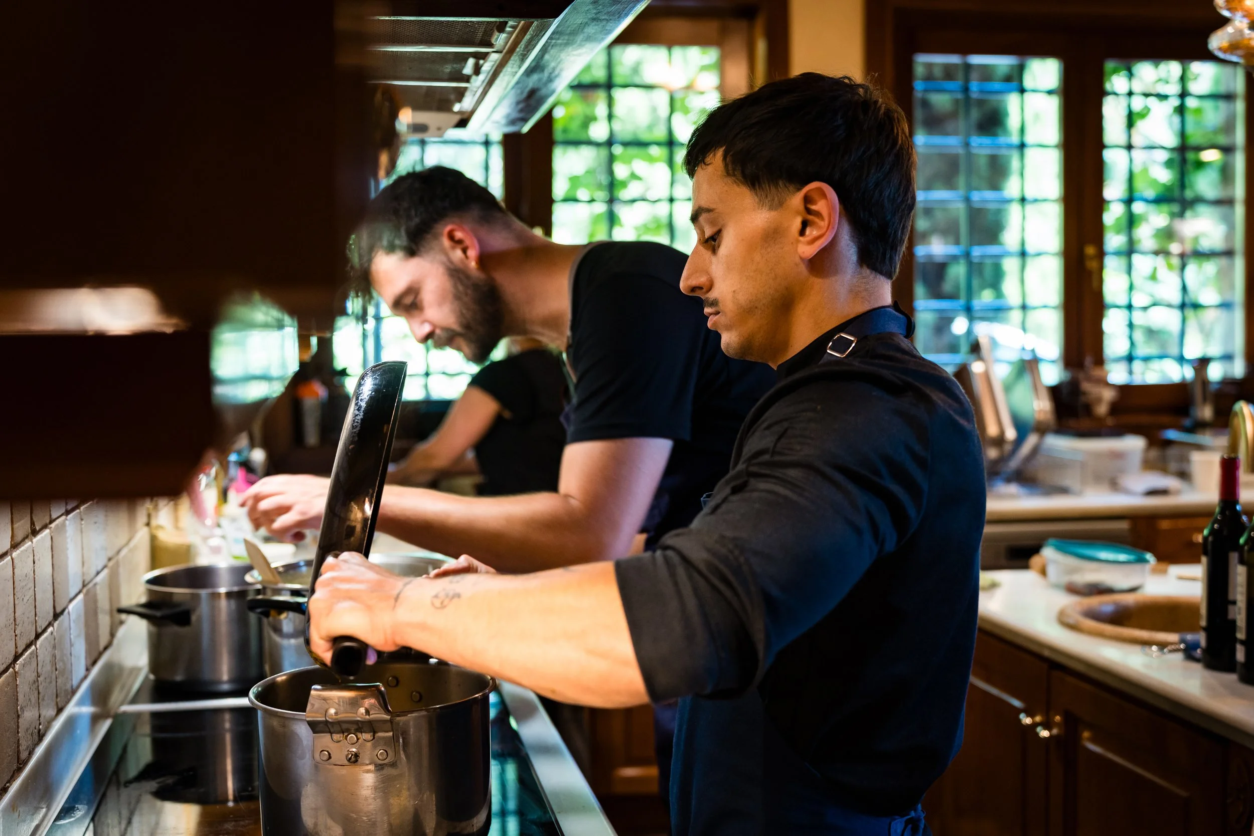 Dos jóvenes cocineros trabajando en la cocina de un restaurante con ventanas grandes y mucha luz natural.