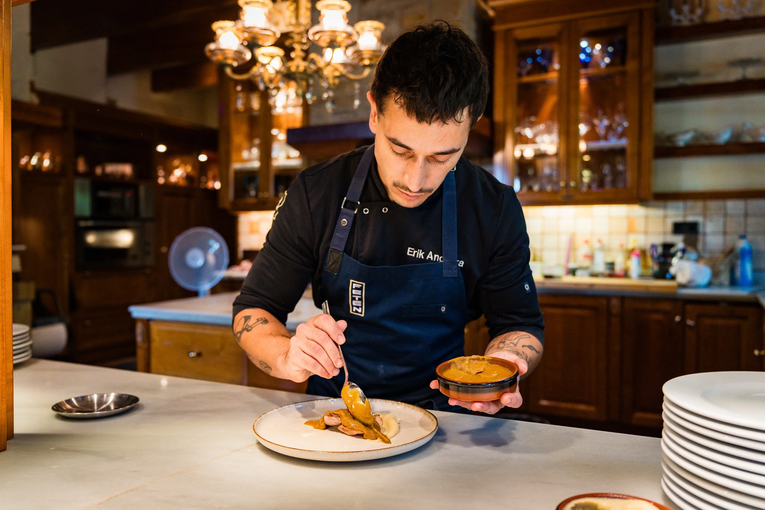 Un hombre con tatuajes y camiseta negra sirviendo comida en un plato, en un ambiente de restaurante con luz cálida y estanterías de madera.
