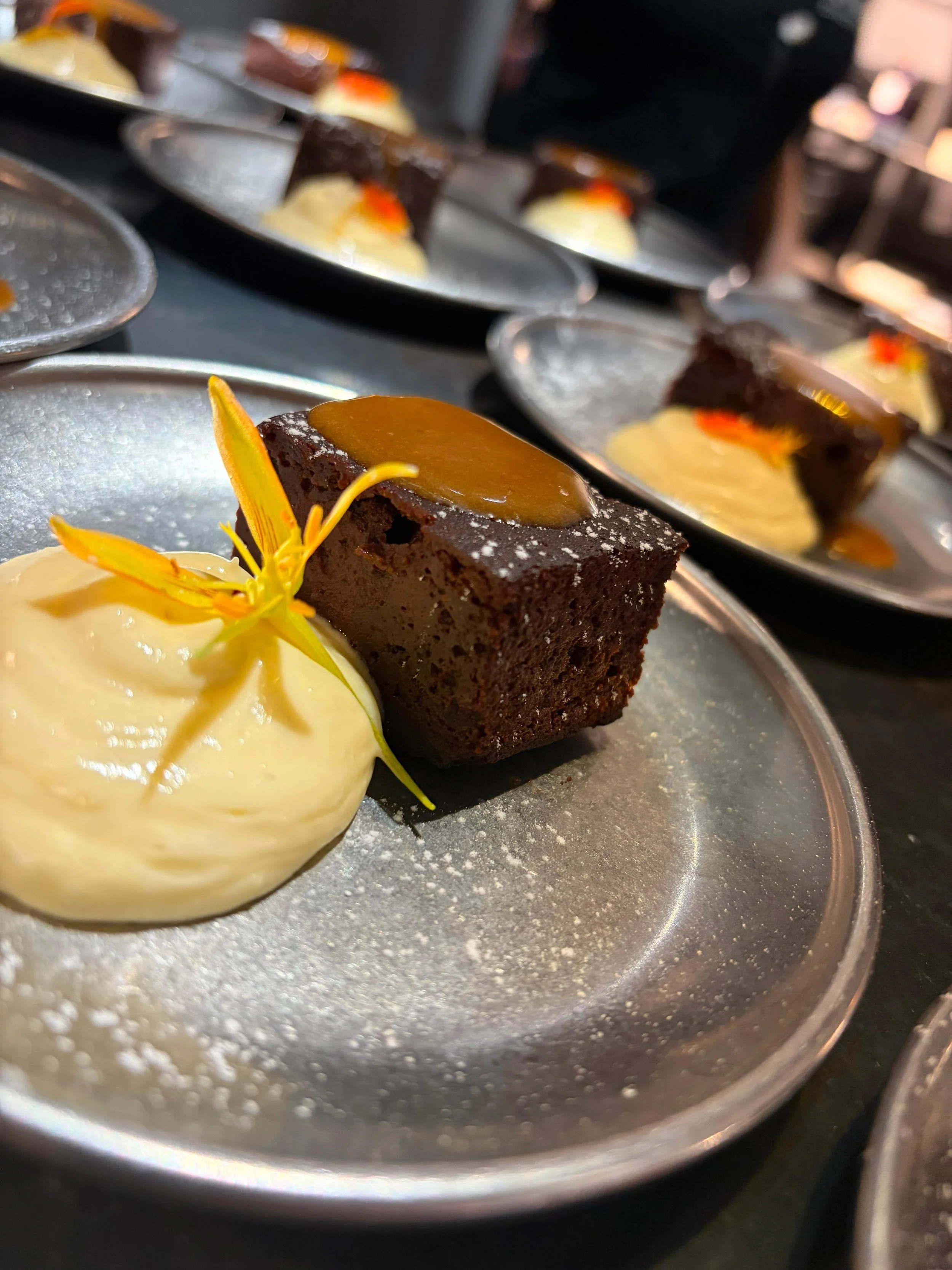 Postre de chocolate acompañado de crema y decorado con una flor amarilla.