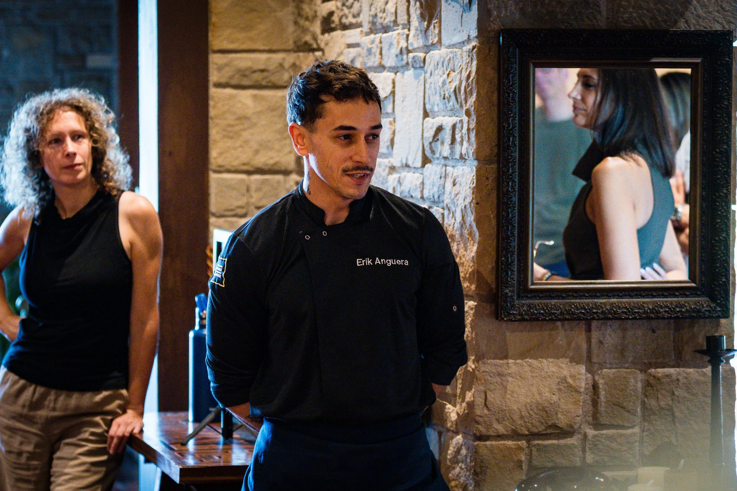 Hombre joven con uniforme negro con el nombre Erik Anguera en un establecimiento de comida o bar, con paredes de piedra y un espejo que refleja la parte superior de su rostro y el ambiente del lugar, acompañado de una mujer con cabello rizado y otra 