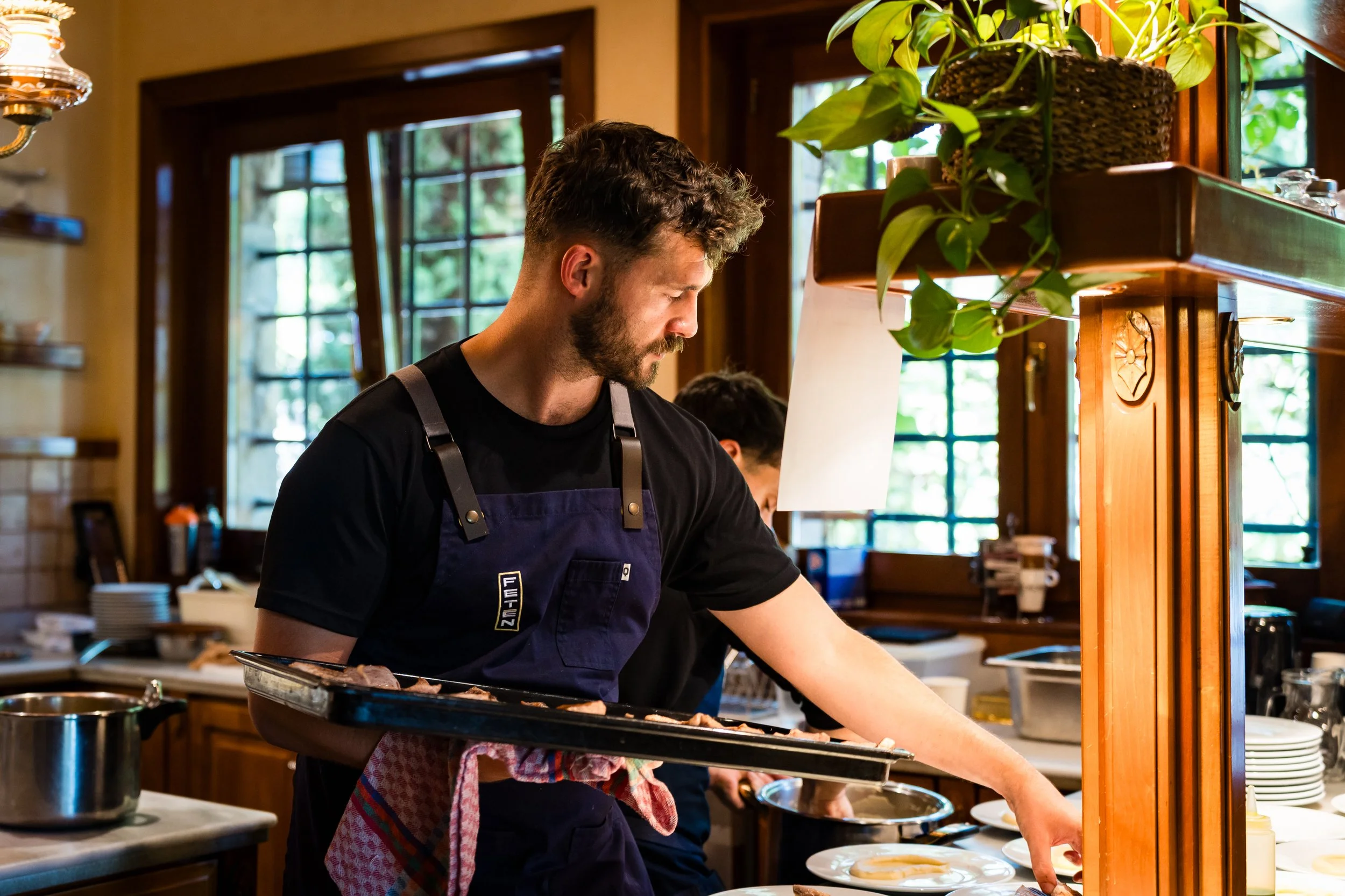 Un hombre con barba y cabello rizado trabajando en la cocina, vistiendo un delantal azul oscuro y sosteniendo una bandeja, en un ambiente con ventanas de madera y plantas decorativas.