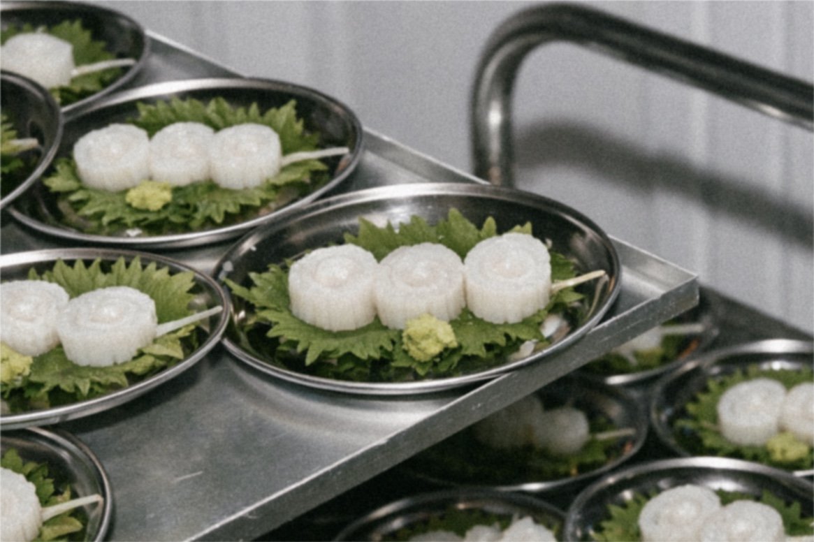Platos de sushi con rollos de arroz y hojas verdes en tazas de metal, en una mesa de trabajo.