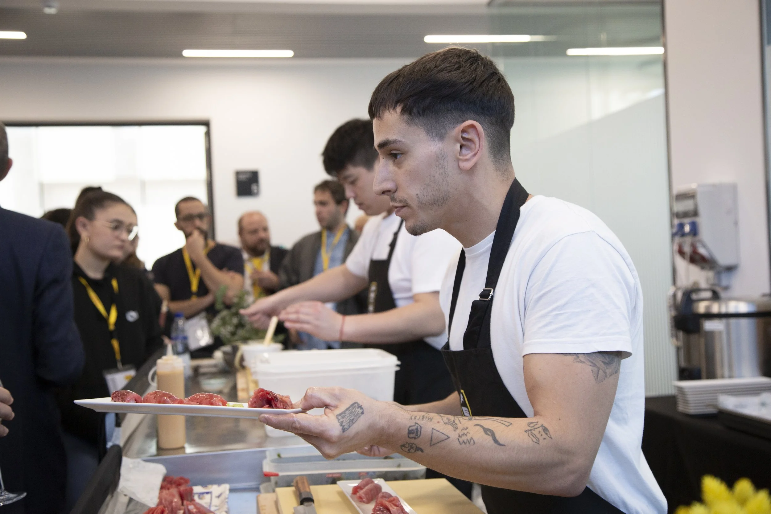 Un joven chef con tatuajes en los brazos sirviendo carne cruda en un plato en una cocina comercial, rodeado de personas que parecen ser participantes en un evento culinario.