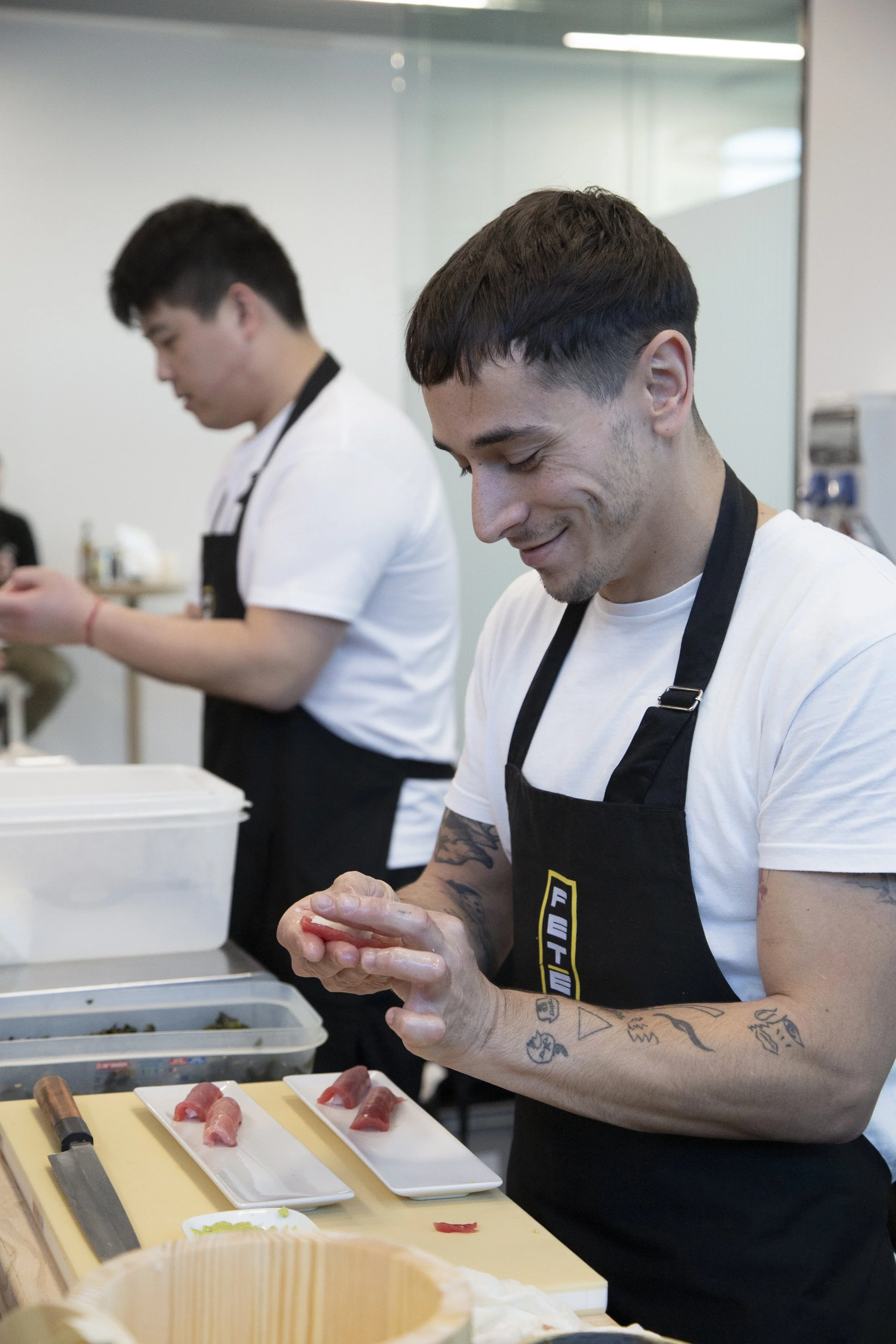 Dos hombres en delantales preparando sushi en una mesa con ingredientes y cuchillos, en un ambiente de cocina o taller de cocina.