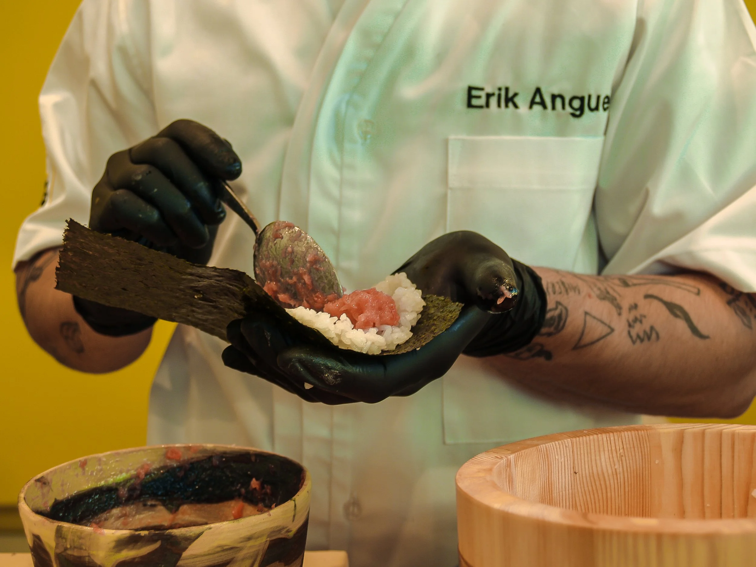 Persona sosteniendo una pieza de sushi en una tabla, con tintas negras en los dedos y una bata blanca con el nombre 'Erik Angu'.