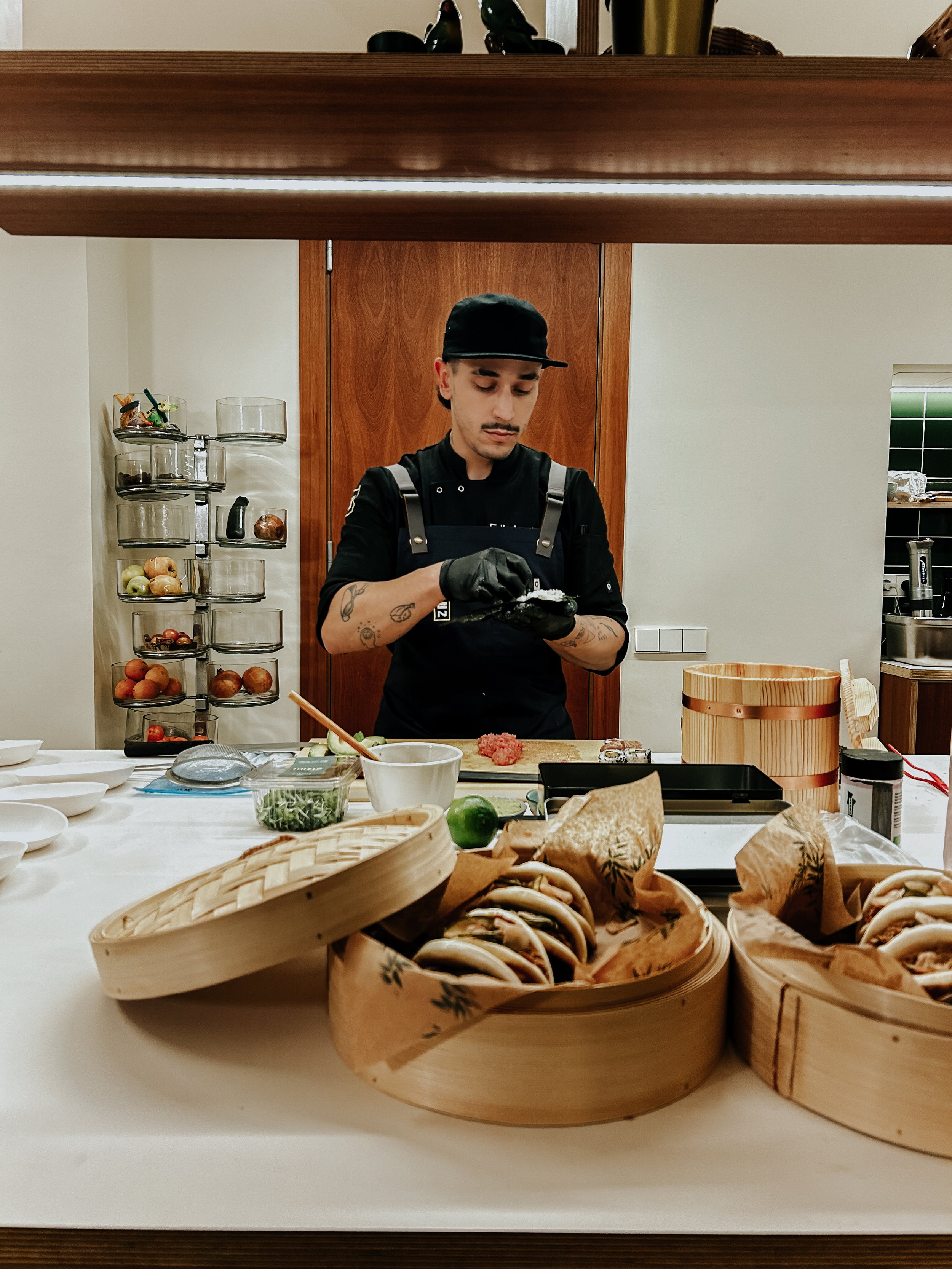 Mesa con bandejas de comida japonesa, sushis, palillos, y ingredientes frescos en una cocina moderna.
