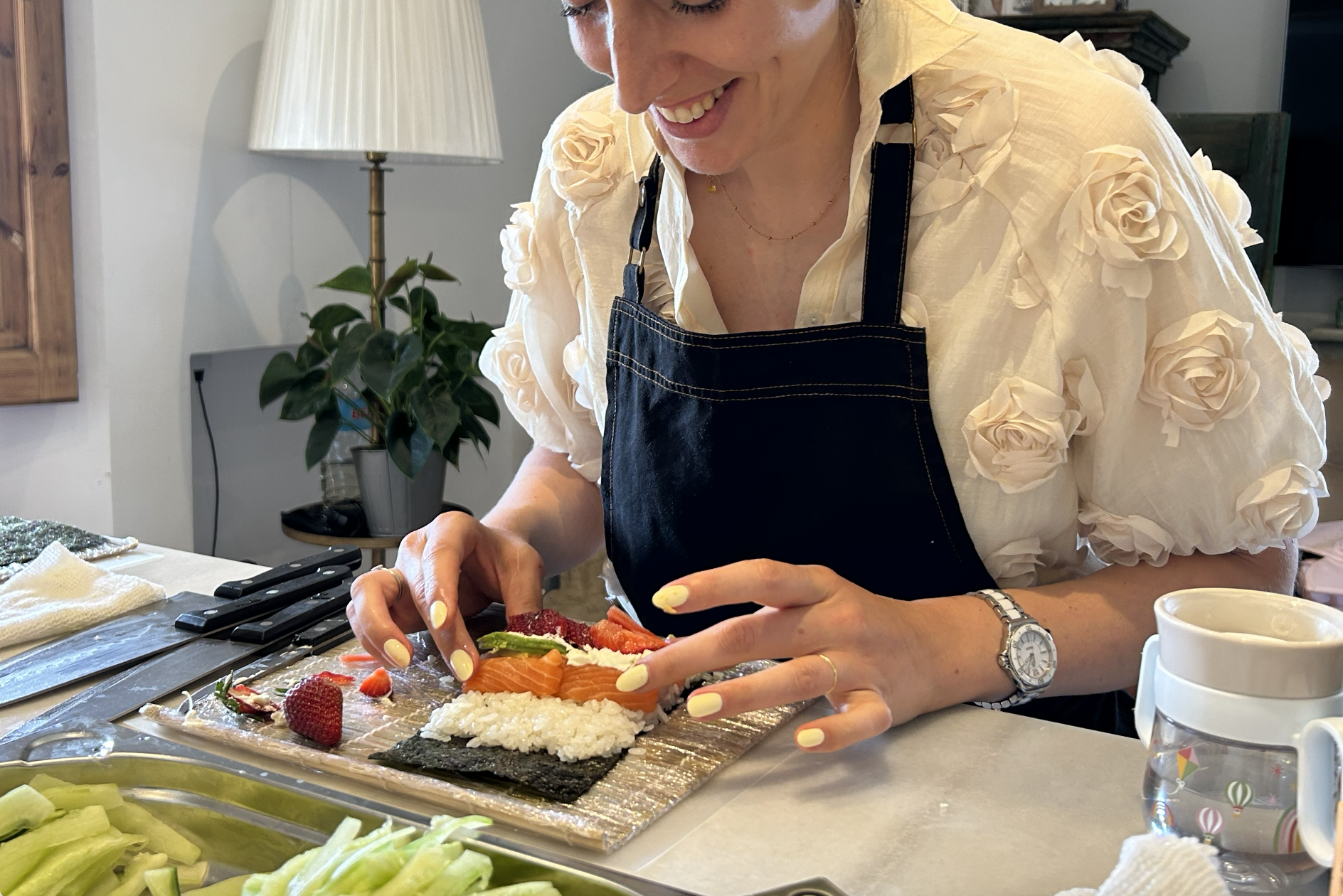 Mujer preparando sushi en una mesa, con ingredientes frescos como pepino y fresa, en un ambiente acogedor con lámpara y planta de interior. Taller de sushi a domicilio.