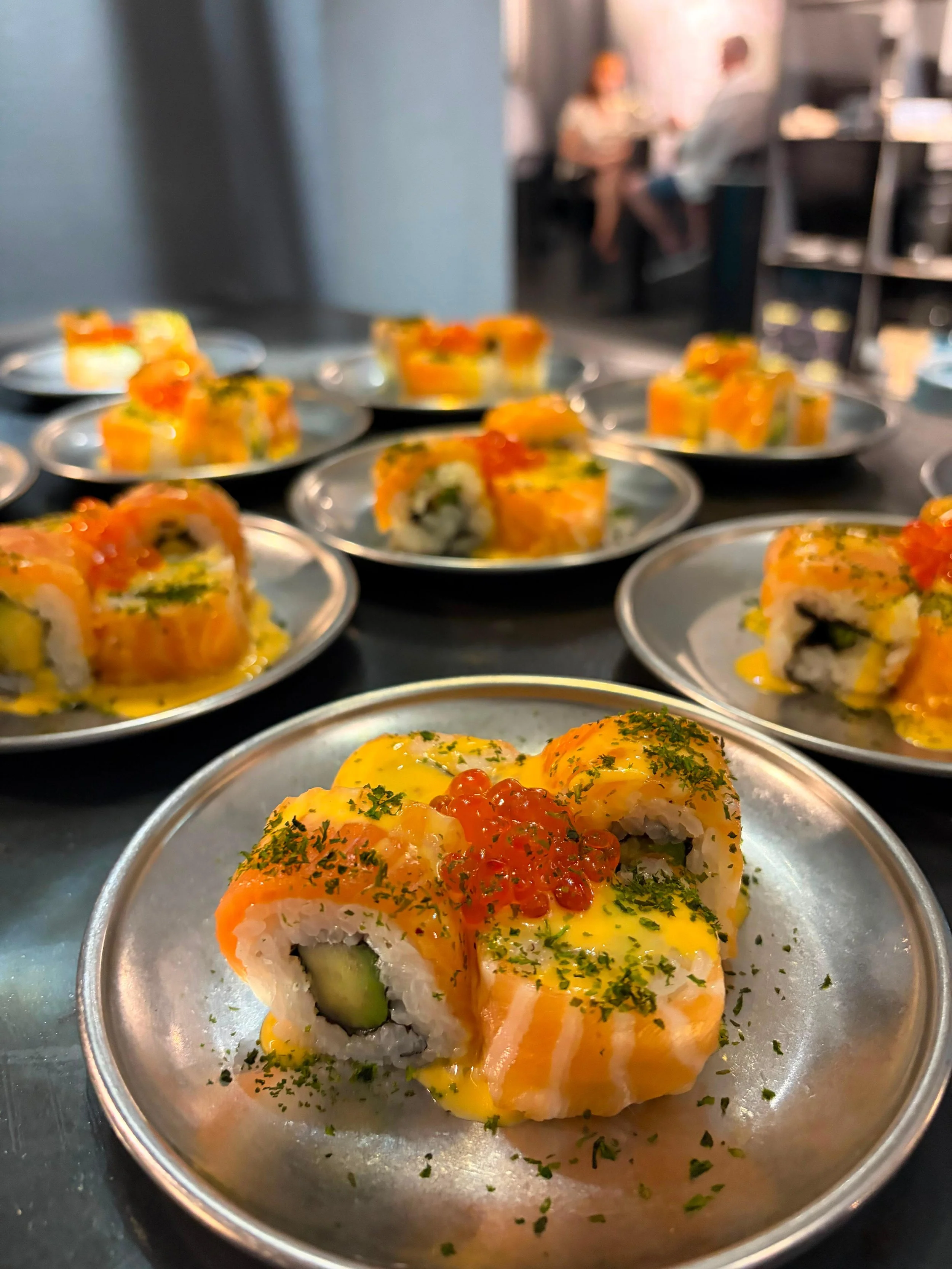 Un plato con sushi enrollado, cubierto con salsa de color naranja y decorado con hierbas verdes y huevas de pescado. En el fondo, otros platos con sushi en un comedor de restaurante.