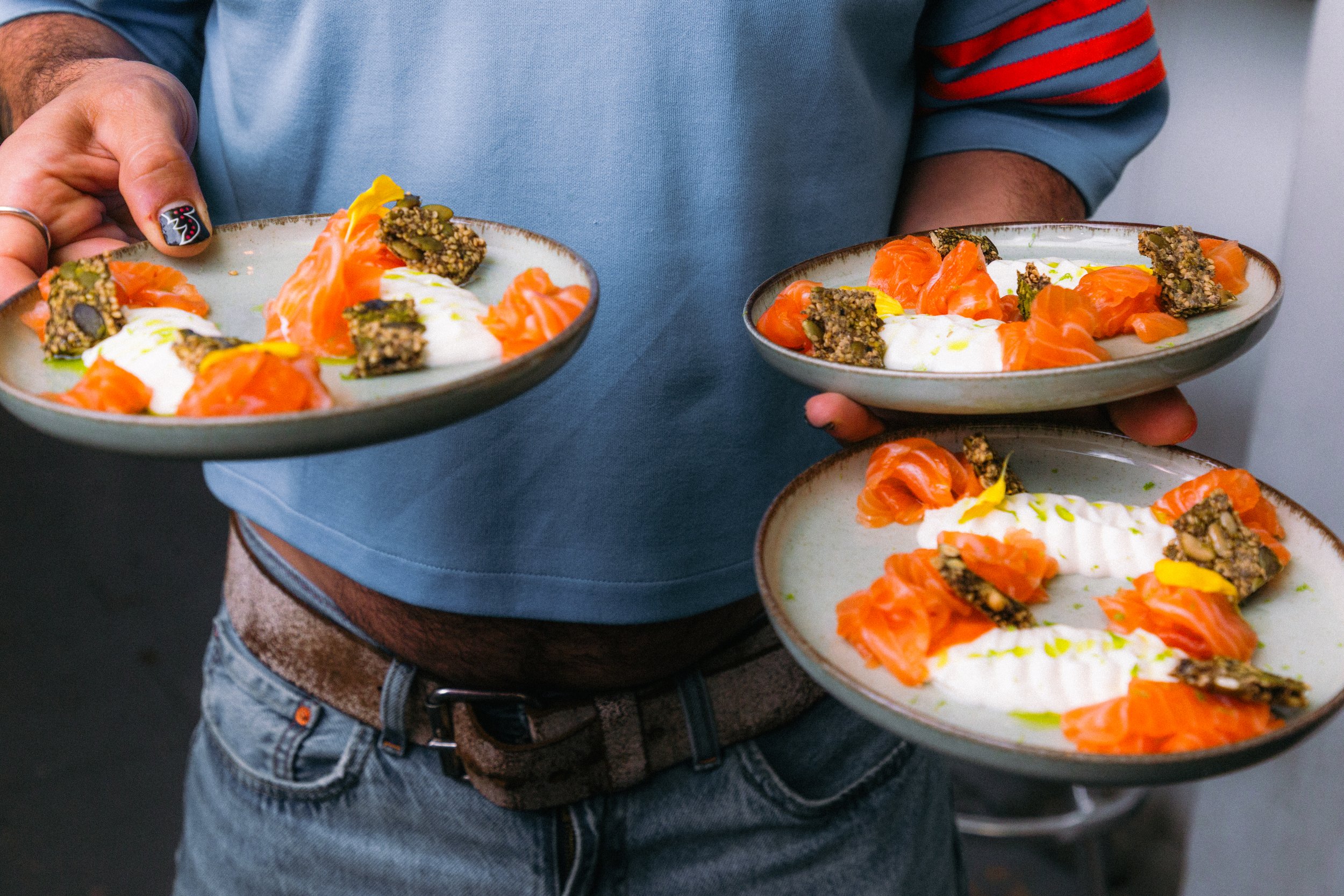 Persona sosteniendo platos con salmón ahumado, queso y semillas, en un entorno interior.