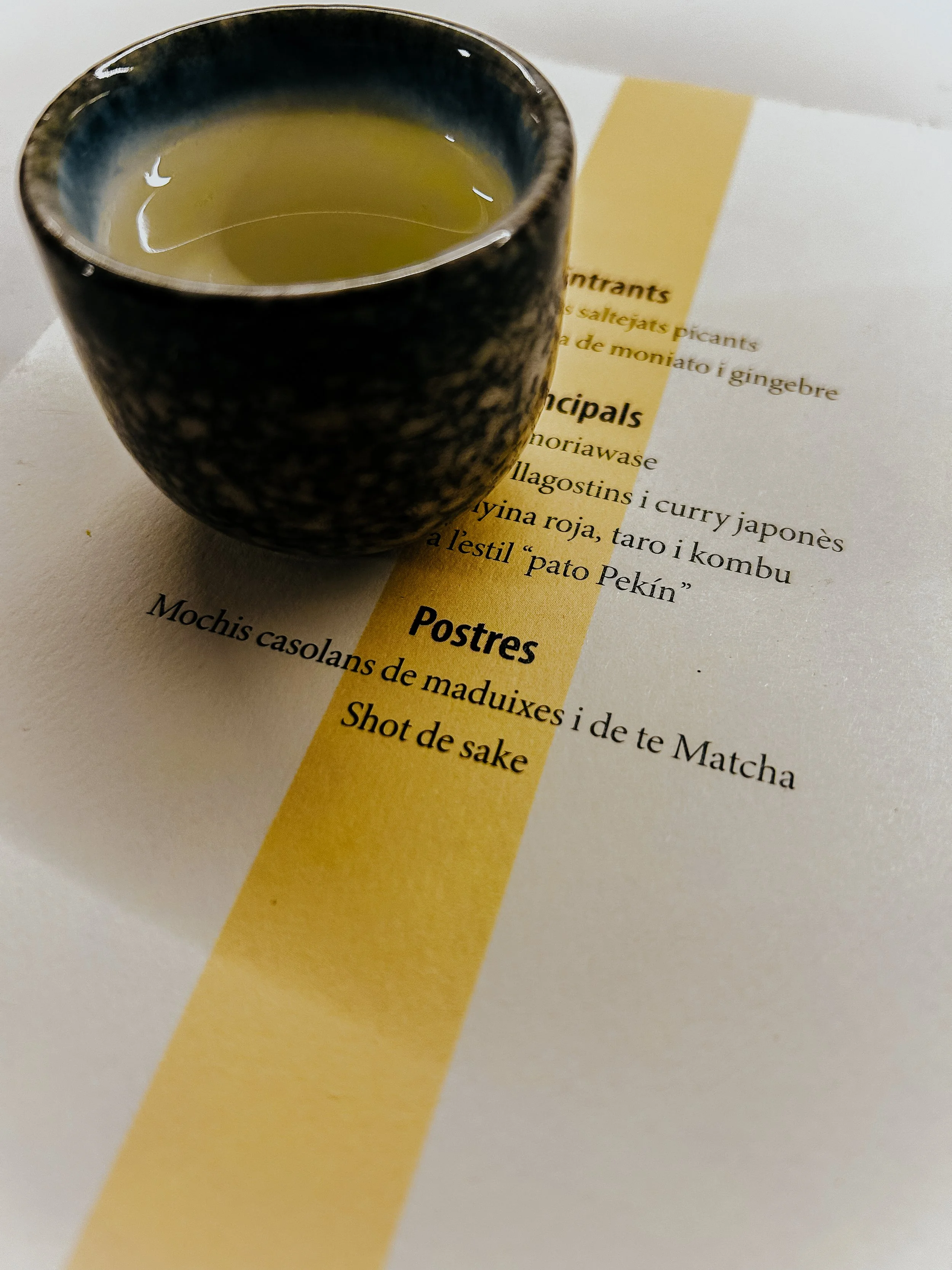 Una taza negra con interior azul y tazón con líquido amarillo, colocado sobre un menú en papel con texto en catalán.