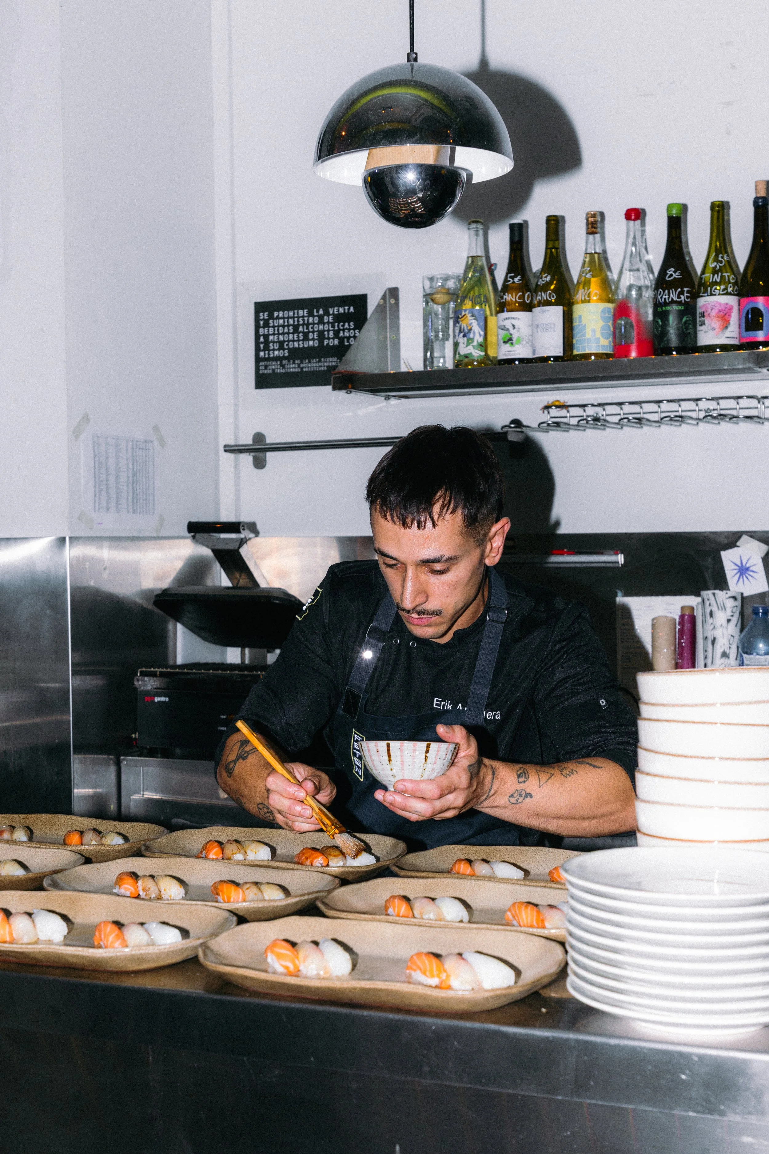 Un hombre con tatuajes decorativos en los brazos y cabello oscuro trabaja en una cocina preparando sushi en bandejas con arroz y salmón, con platos apilados blancos en primer plano y una estantería con botellas de bebidas en el fondo. Atún rojo.